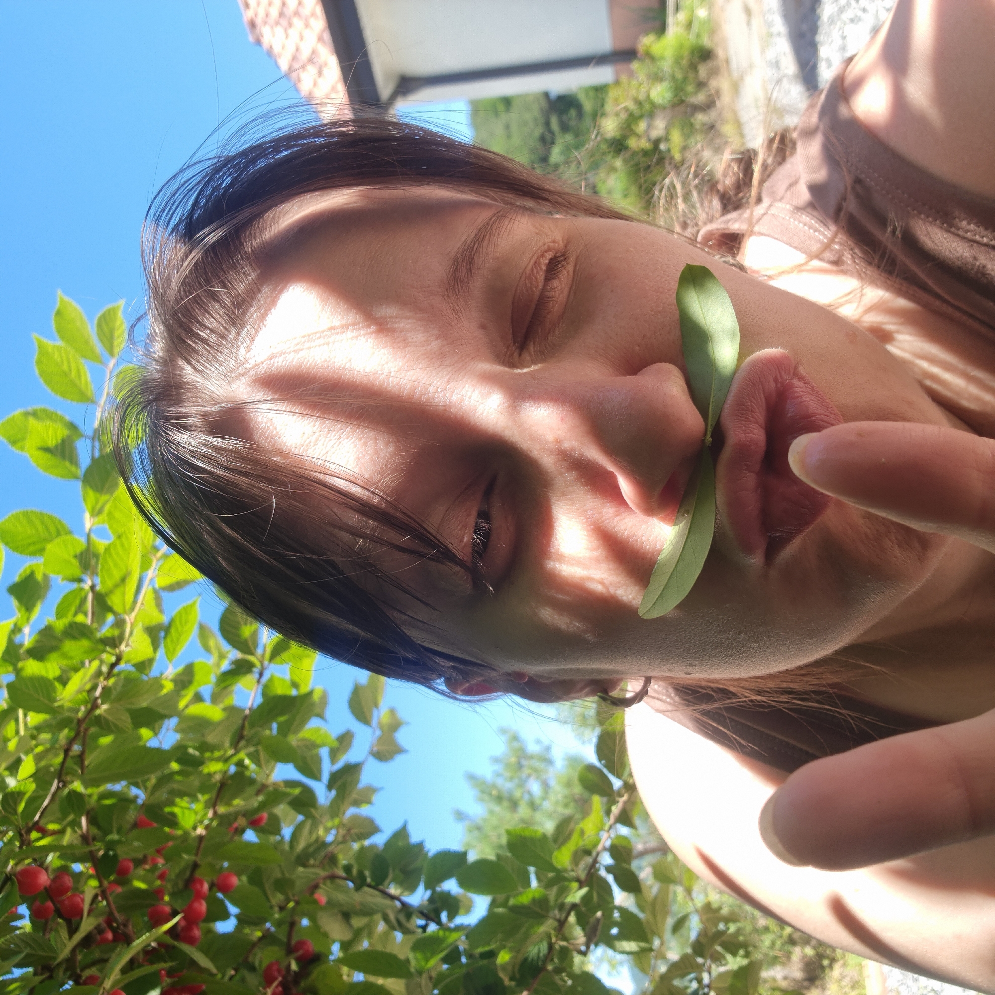 A selfie-style, close-up shot shows a person's face, positioned centrally. The person has dark brown hair, and some strands fall across their forehead. They are looking at the camera with one eye open and the other closed, with their lips pursed. A green leaf is placed horizontally above their upper lip, resembling a mustache. Sunlight casts dappled shadows across the person's face. Their right hand is visible in the foreground, with the index finger extended towards the camera.
The background is blurred but shows greenery, including leaves and red berries on a branch in the upper left quadrant. The sky is a clear blue. In the upper right, a portion of a building with a tiled roof is visible, and further in the background, more foliage and a hillside can be seen. The scene appears to be outdoors, possibly in a garden or natural setting. The overall impression is one of playful engagement with nature.