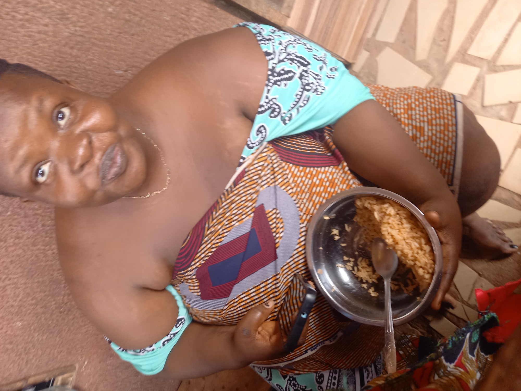 A close-up, high-angle shot captures a person, likely a woman, with dark skin, sitting or reclining in Onitsha, Nigeria. She looks towards the camera with an animated expression, her mouth slightly open and eyes wide, suggesting surprise or engagement. She wears a vibrant, multi-patterned traditional garment; the sleeveless upper portion is light blue with black and white designs, while the main body features orange, brown, red, and grey geometric patterns. A thin gold chain is visible around her neck.
She holds a silver-colored metal bowl containing light-colored rice with a spoon resting inside. Her left hand is seen holding a black smartphone against her body, while her right hand supports the bowl of food. The setting appears casual, with a textured floor of light brown and tan square and rectangular tiles visible in the background. Part of a bare foot with dark nail polish is also visible in the lower right, possibly belonging to the subject or another person nearby. The scene conveys a moment of eating or sharing food in a relaxed, daytime environment. No discernible text is visible in the image.