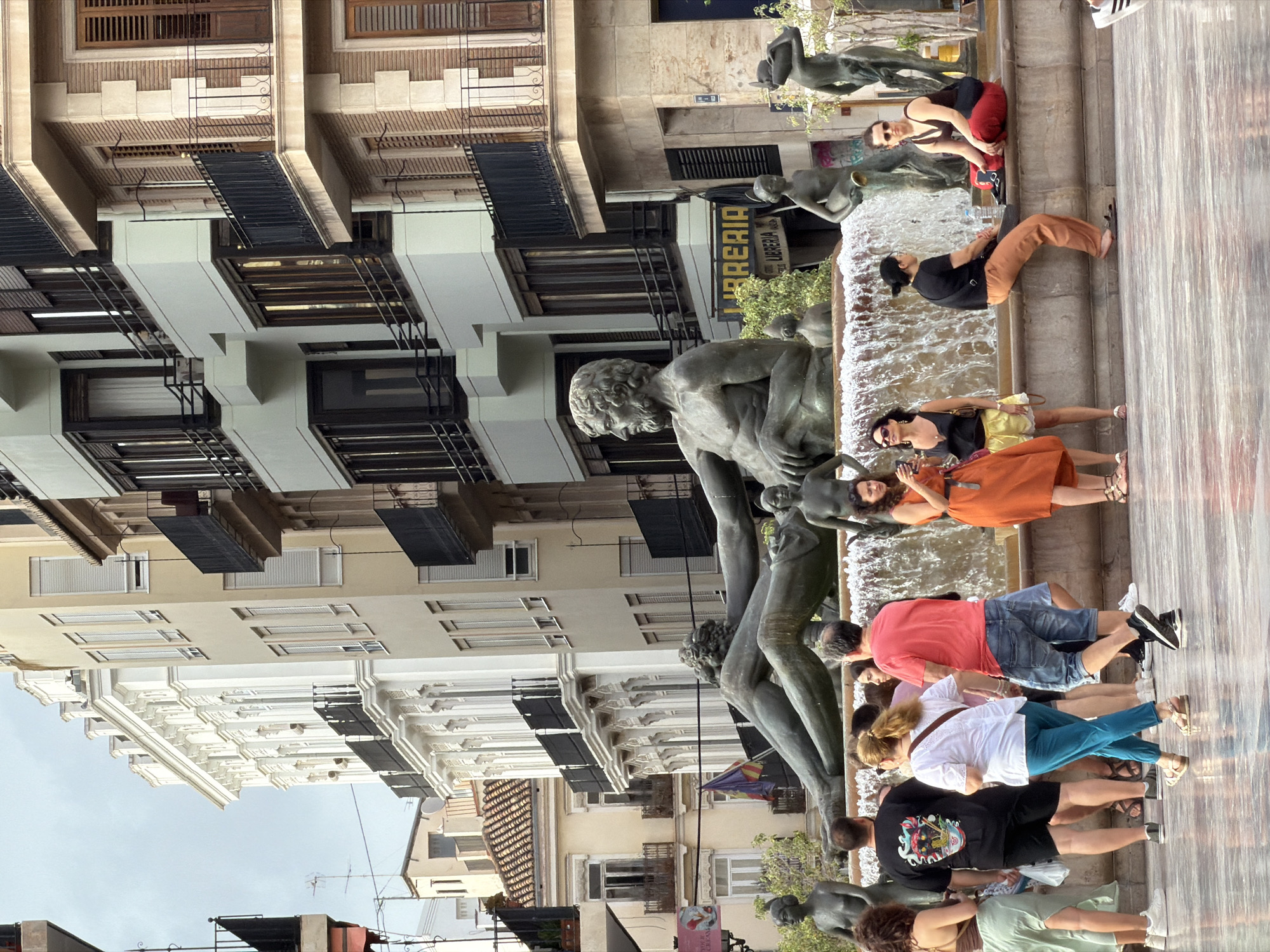 The image depicts a busy public square with a large fountain as the central focus. The fountain features a monumental bronze sculpture of reclining figures, with several smaller bronze statues of nymphs and cherubs around it. Water cascades from the fountain, creating a mist that catches the light.
In the foreground, several people are gathered around the fountain. On the left, a woman with curly brown hair wears a light green skirt and a brown top. Next to her, a man in a black t-shirt with a graphic print and black shorts stands with his back to the camera. To his right, a woman in a white shirt and turquoise pants walks away, while a man in a red t-shirt and blue shorts walks in the same direction.
Further to the right, a woman in an orange dress stands near the fountain, holding a smartphone. A woman in a black top and yellow shorts is seated on the edge of the fountain. To her right, a woman in a black tank top and red pants sits on the fountain's edge, looking to her left. Another woman in a black shirt and brown pants is also seated on the fountain's edge, further to the right.
The background is dominated by elegant multi-story buildings with balconies and shutters. The architecture suggests a European city, with ornate detailing on some of the facades. A flag, likely a city or regional flag, is visible hanging from one of the buildings. The sky is overcast, contributing to a diffused lighting across the scene. The ground in front of the fountain appears to be a polished stone or tile, reflecting the surrounding elements.