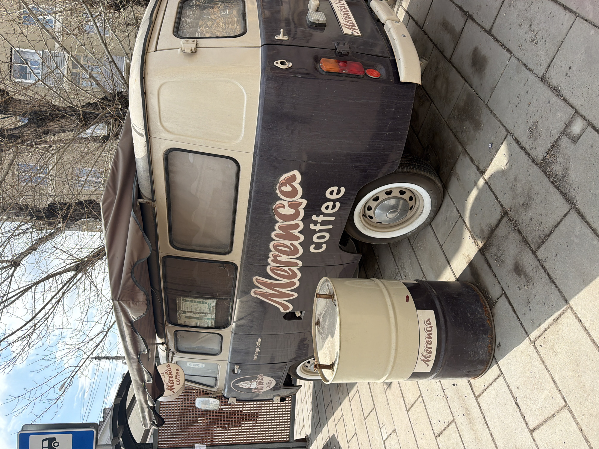 A coffee van called "Merenca coffee" parked on a brick sidewalk. The van is painted cream and dark gray with the words "Merenca coffee" written on the side and a logo. A metal barrel painted to match the van is next to it, also with "Merenca" written on it. Trees are visible in the background along with a building and a charging station. The photograph was taken in Krasnoyarsk, Russia.