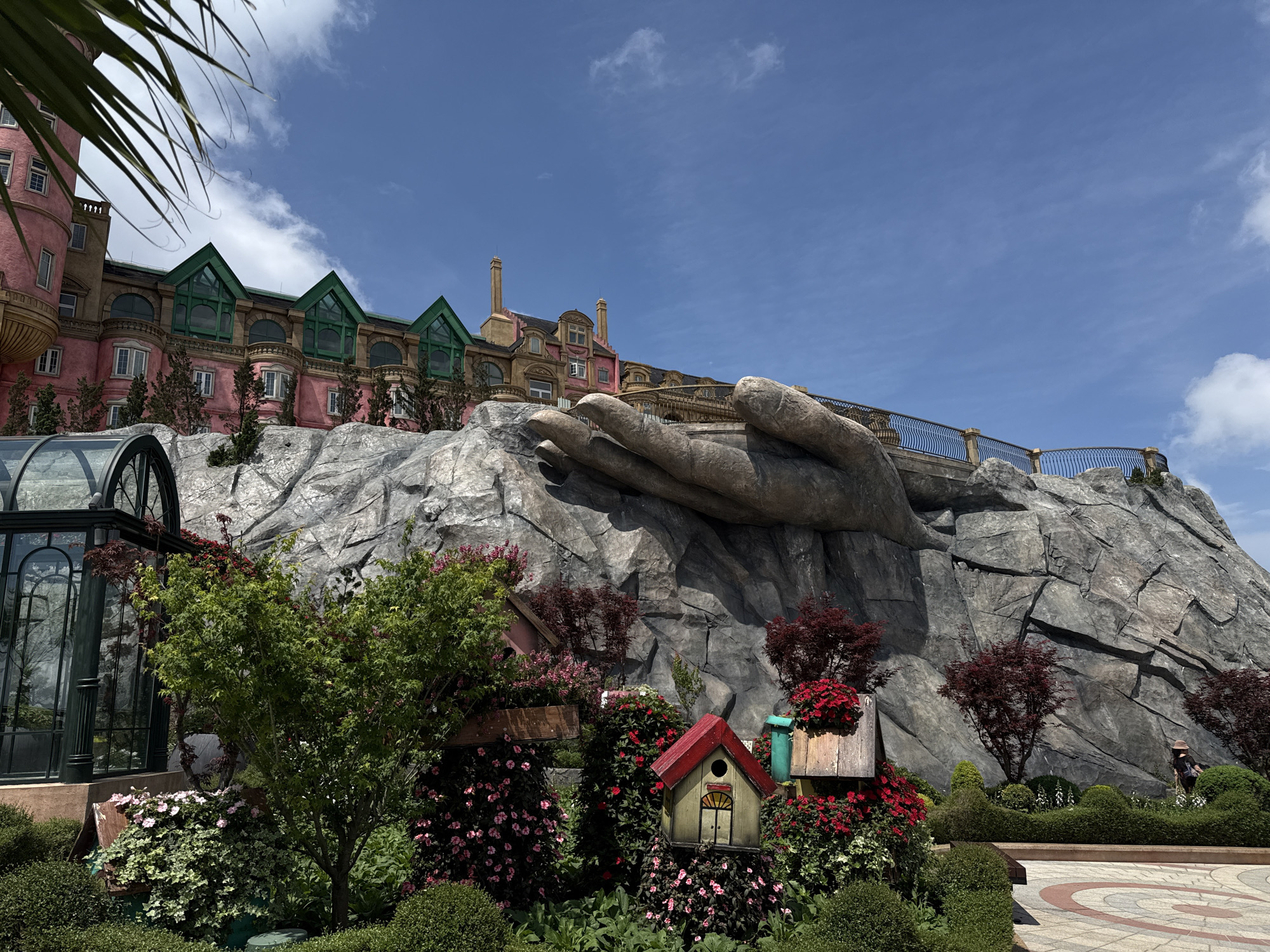 The image shows a building complex at the Ba Na Hills resort in Đà Nẵng, Vietnam. A pink multi-story building with green roofs is visible atop a gray, rocky structure that appears to be sculpted into the shape of giant hands. A decorative metal railing runs along the top edge of the hand-shaped rocks. Below, there is a garden area with lush greenery, flowers, decorative birdhouses, and a greenhouse-like structure on the left. The sky is blue with some clouds. A person wearing a hat is partially visible in the background near the right edge of the image.
