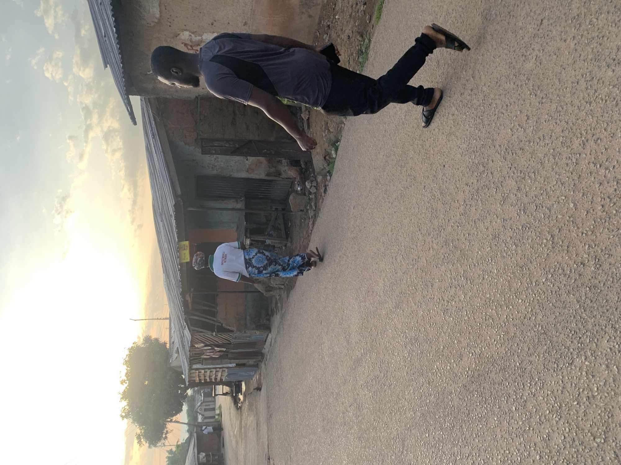 The image depicts a street scene in Jos, Nigeria, during what appears to be either sunrise or sunset, given the warm lighting and soft clouds. In the foreground, a man with dark skin and a beard is walking away from the camera towards the right side of the frame. He is wearing a dark grey t-shirt with a black diagonal design on the chest, black pants, and black sandals. He is holding a dark object, possibly a phone, in his right hand.
Further ahead on the street, also walking away from the camera, is a person wearing a white t-shirt with green trim around the neck and the text "IMPACTWORLD NETWORK" printed on the back in green. This person is also wearing a long, colorful patterned skirt or wrap.
The environment consists of a paved road with a gravelly texture. Along the left side of the road, there are several single-story structures with corrugated metal roofs and walls constructed from mud or rough materials. These buildings appear to be shops or residences, some with open fronts displaying various items or structures within. A large tree with lush green foliage is visible on the left side of the frame, behind some of the buildings. The sky above is bright with scattered clouds, suggesting natural light as the primary illumination. The overall impression is of a somewhat rustic urban or semi-urban setting.