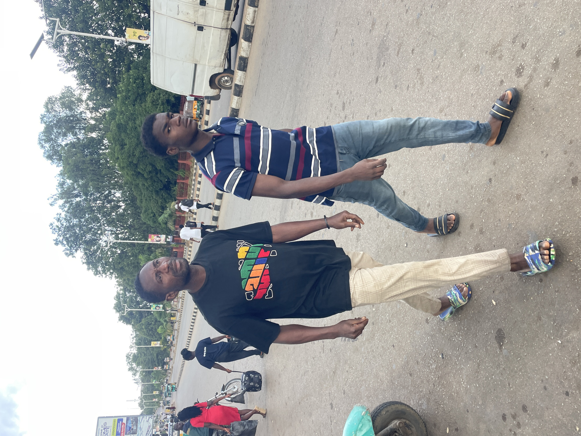 Two men are walking side-by-side on a street in Jos, Nigeria. The man on the left is wearing a black t-shirt with a colorful graphic and light-colored plaid pants. He has dark skin and a serious expression. The man on the right is wearing a blue and red striped polo shirt and blue jeans. He is looking off to the side. In the background, there are trees, streetlights, a white van, and other people walking. The scene appears to be a busy street with traffic.