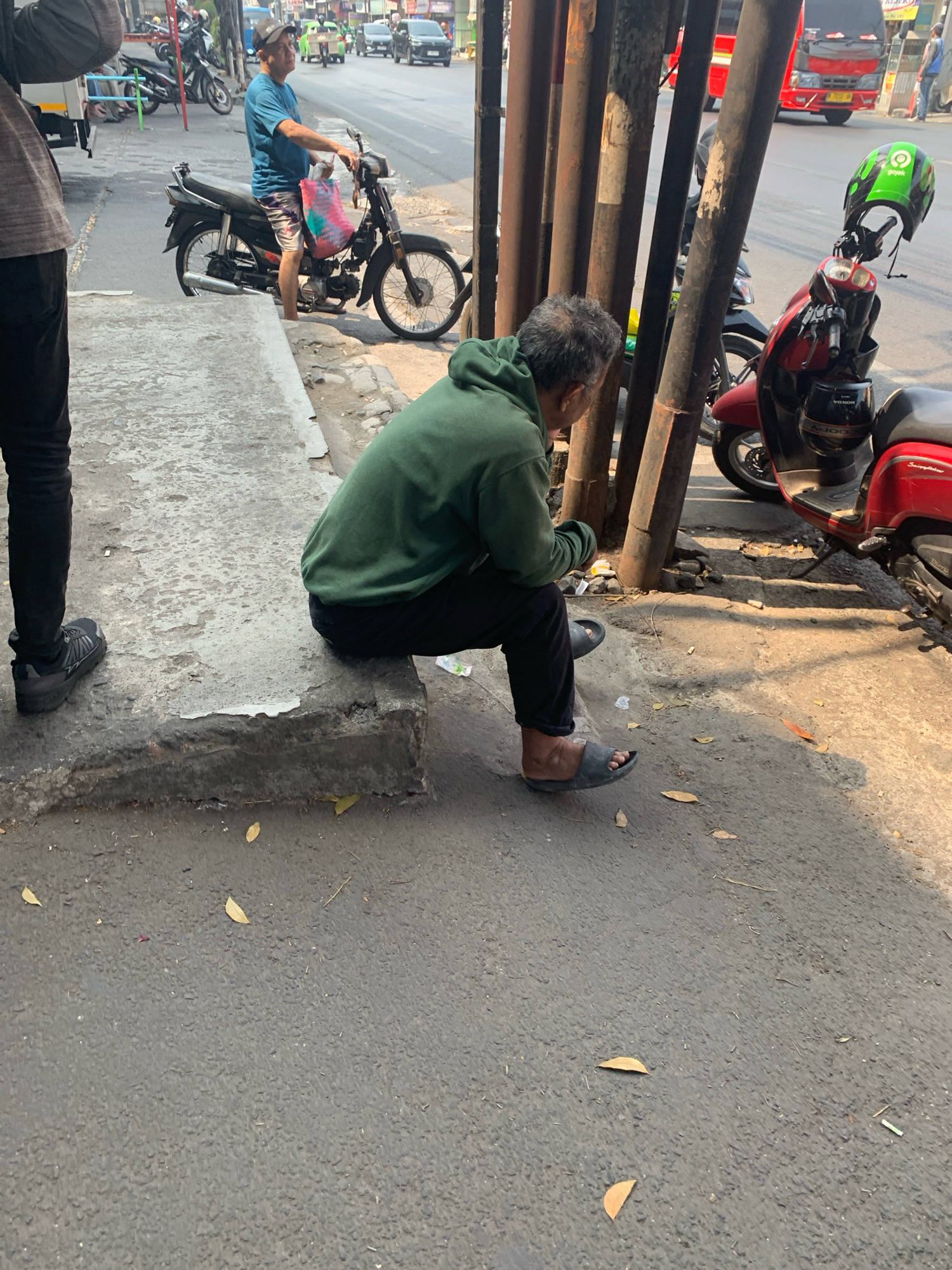 The image captures a street scene in Malang, Indonesia, likely during the day, given the bright sunlight casting shadows. In the foreground, a man wearing a green hooded sweatshirt and black pants sits on a concrete curb. He is facing away from the camera, looking down at his hands. He is wearing dark grey flip-flops. To his left, another person, partially visible, stands wearing black pants and dark sneakers.
Behind the seated man, a red scooter is parked, with a green helmet with "Gojek" branding attached to the handlebars. Further back, a black motorcycle is also parked. The background shows a street with several vehicles, including cars and a red truck. Buildings and trees line the street, suggesting an urban or semi-urban environment. The ground is paved with asphalt and concrete, with fallen leaves scattered around. The overall impression is of a casual moment on a busy street in Malang.