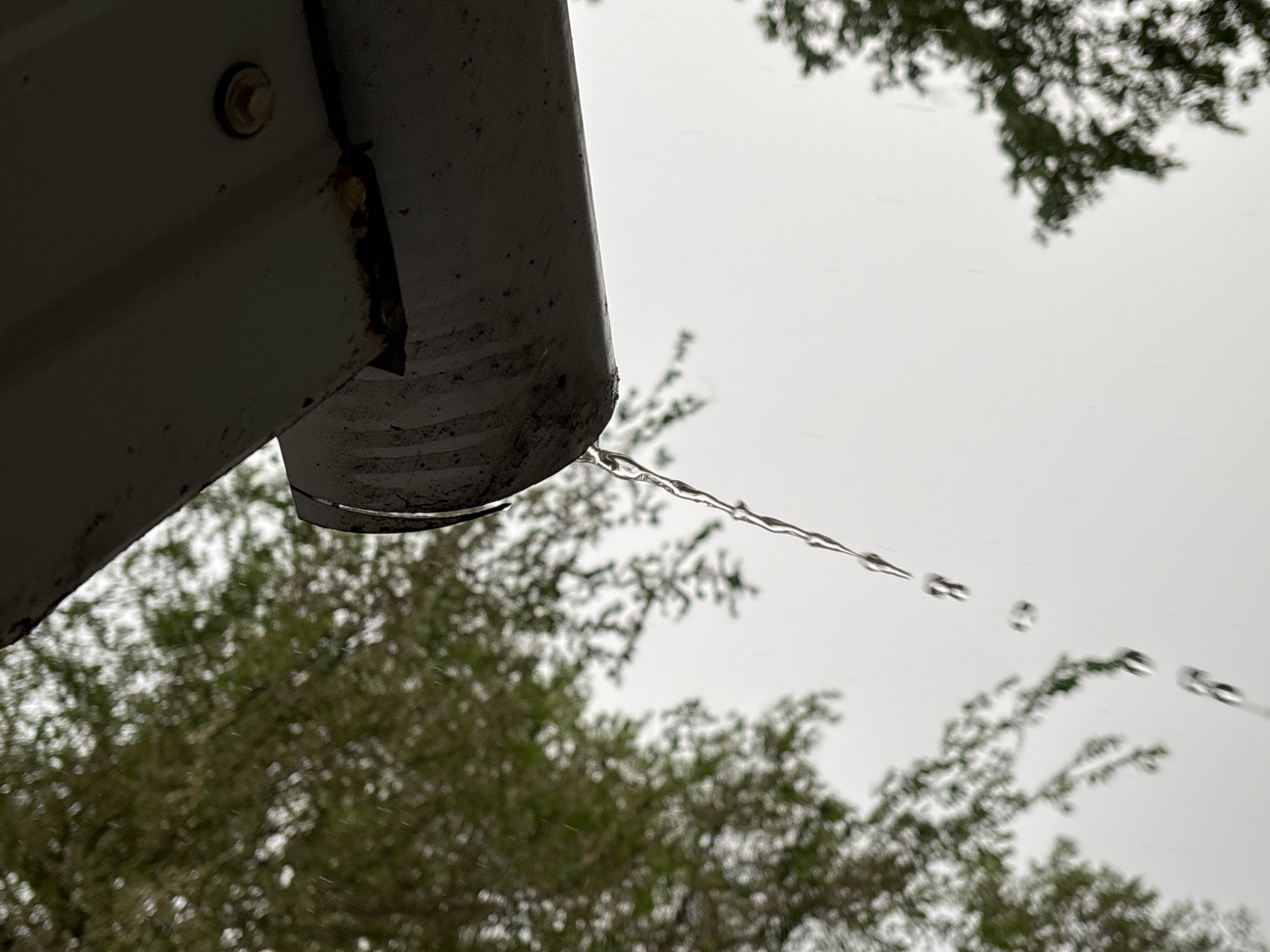 A close-up, low-angle shot depicts a metal gutter, likely part of a building's drainage system. Water droplets are visible in mid-air, trailing off the edge of the gutter, suggesting recent or ongoing rain. Background features blurred foliage and a gray, overcast sky. There are no people visible in the image. The city is unknown.