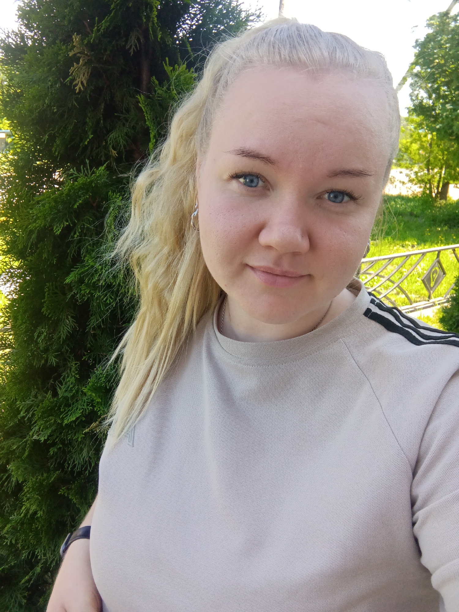 A fair-skinned woman with light blonde hair pulled back in a ponytail is the central subject of this medium-shot outdoor photograph. Her eyes are blue, and she wears a beige, long-sleeved shirt with three black stripes on the shoulders. She has small silver hoop earrings.
Behind the woman on the left is a dense, green bush or shrub. In the background, slightly out of focus, is a green grassy area with trees and a metal fence. The photograph was taken in Фоки, Russia.