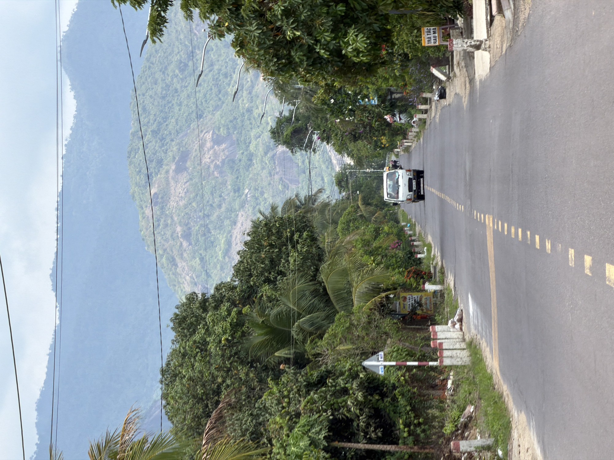 The image shows a straight, paved road in what appears to be a rural area of Diên Thọ, Vietnam. The road is lined with trees and vegetation on both sides, and in the background, there are rolling, mist-covered mountains.
In the foreground, a white van is driving away from the camera down the center of the road. The road is marked with a dashed yellow line in the center and a solid yellow line on the right side. On the left side of the road, there are several white posts with red tops, marking the edge of the pavement. A white and red striped signpost is visible on the left.
The vegetation consists of dense green trees, including palm trees, and some flowering bushes with red and pink blooms. On the right side of the road, a yellow sign with Vietnamese text is partially visible. The sky is overcast. The scene suggests a peaceful, natural environment with limited human activity, primarily focused on the road and the approaching van.