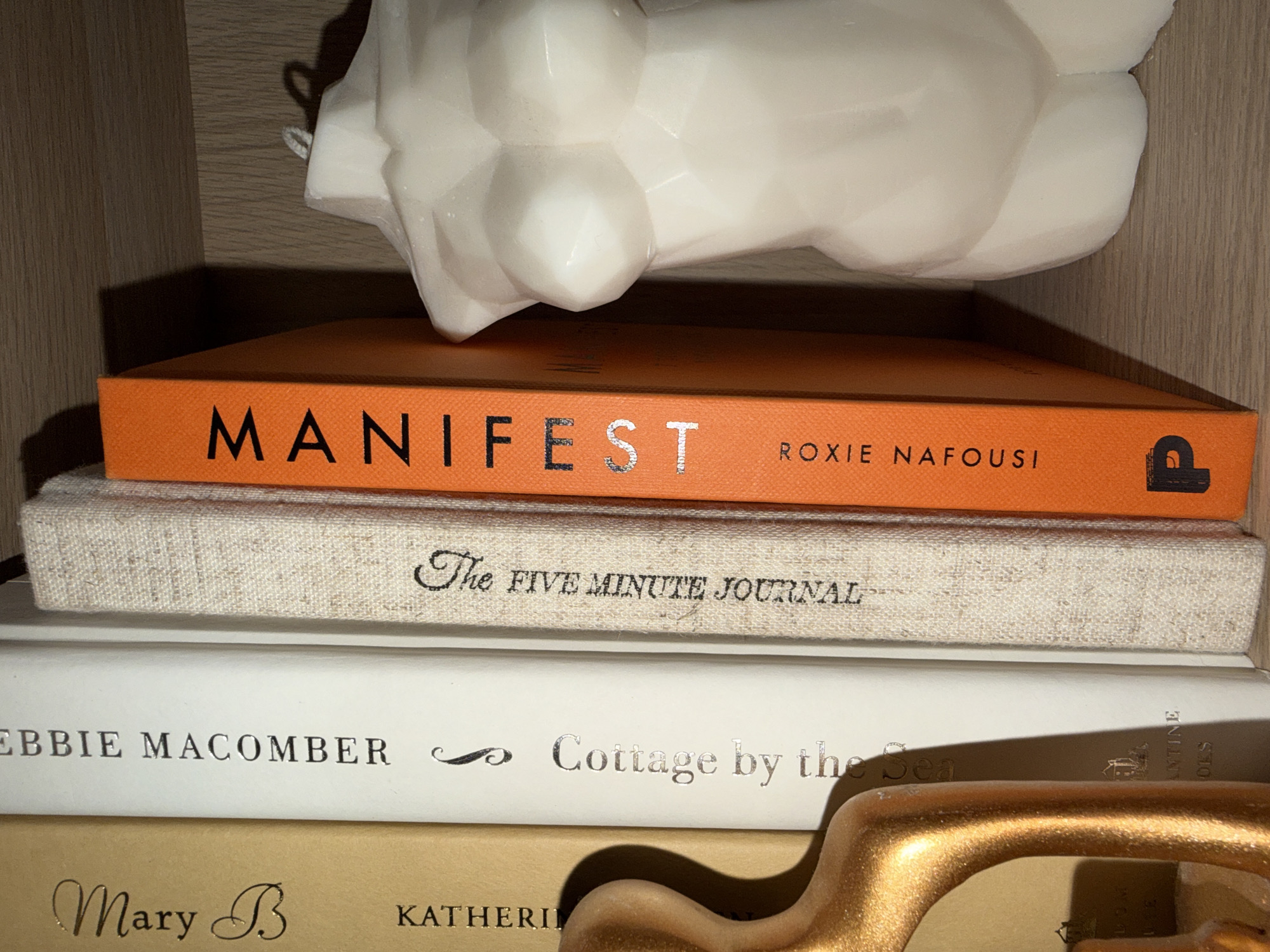 This is a close-up, eye-level, horizontal shot of a stack of books on a wooden shelf. The top book is bright orange and has the title "MANIFEST" in large, metallic silver letters, followed by "ROXIE NAFOUSI" and a small logo. Below it is a cream-colored book with the title "The FIVE MINUTE JOURNAL" in a script font. The next book is white with silver lettering that reads "EBBIE MACOMBER" and then "Cottage by the Sea." The bottom visible book is light brown and has "Mary B" in a script font, and "KATHERI" in silver lettering. A decorative, gold-colored object, possibly a drawer pull or a sculpture, is in the foreground, casting a shadow. At the very top of the image, part of a white, sculpted object, possibly a bust or abstract art piece, is visible. The lighting suggests an indoor setting, likely a home or library, with the light source coming from above and slightly to the right, creating distinct shadows. There are no people or overt indications of a specific time of day or weather.
