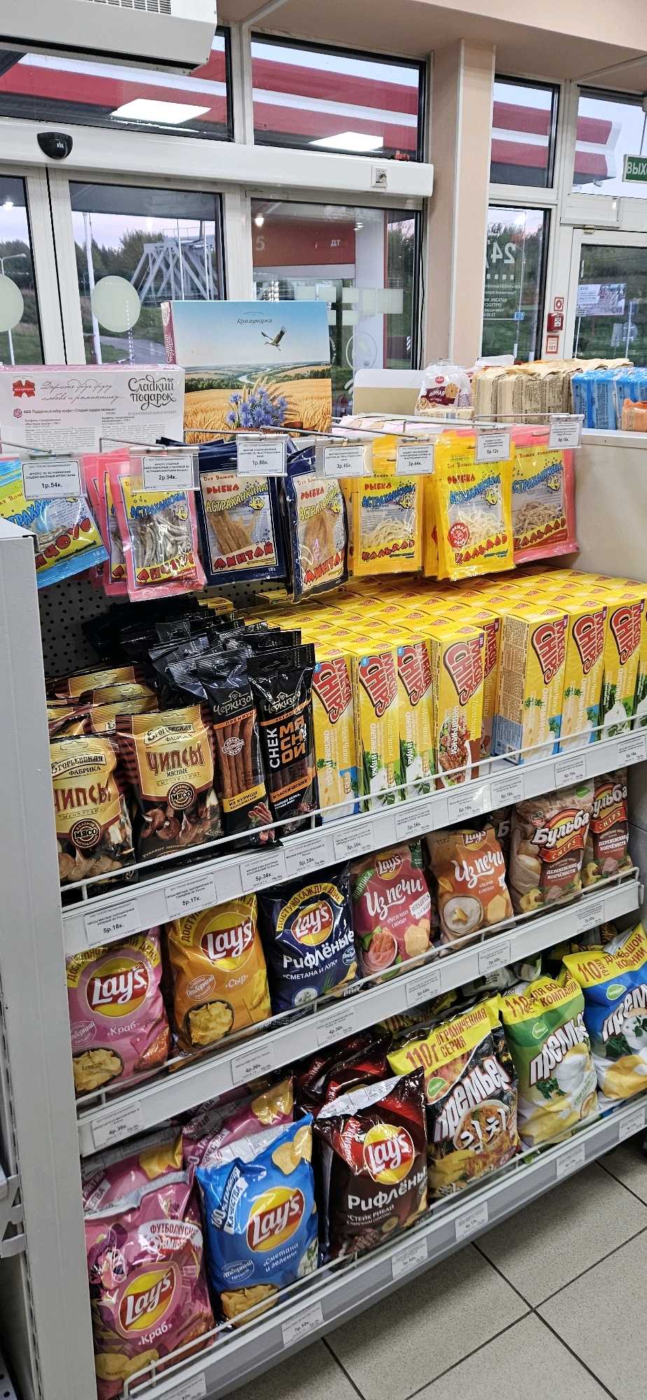 The image shows a well-lit interior view of a store, likely a convenience store or small supermarket, with shelves stocked with various snack items. The products are predominantly packaged chips and dried fish snacks, with some larger boxes of snacks on the lower shelves. The shelves are organized with price tags displayed. The overall environment suggests a commercial retail setting. The specific location in Vitsebsk, Belarus is not determinable from the visual information provided in the image. There are no people or discernible actions captured in the photograph. The focus is entirely on the product display.