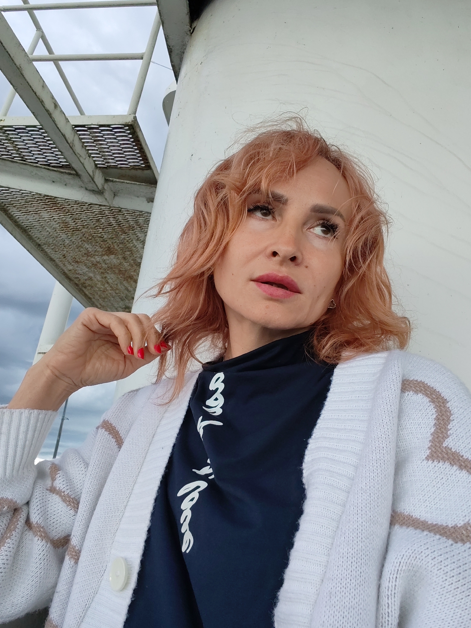 In the city of Shcheleiki, Russia, an image captures a medium shot of a woman with shoulder-length, wavy, peach-colored hair, looking up and to her right. She is wearing a dark blue t-shirt with white script on it, layered under a white knitted cardigan adorned with a beige striped pattern. Her left hand, with bright red nail polish, is raised to touch her hair. The background includes a white cylindrical structure and a metallic gray staircase with perforated treads, suggesting an outdoor industrial or maritime setting. The sky appears overcast.