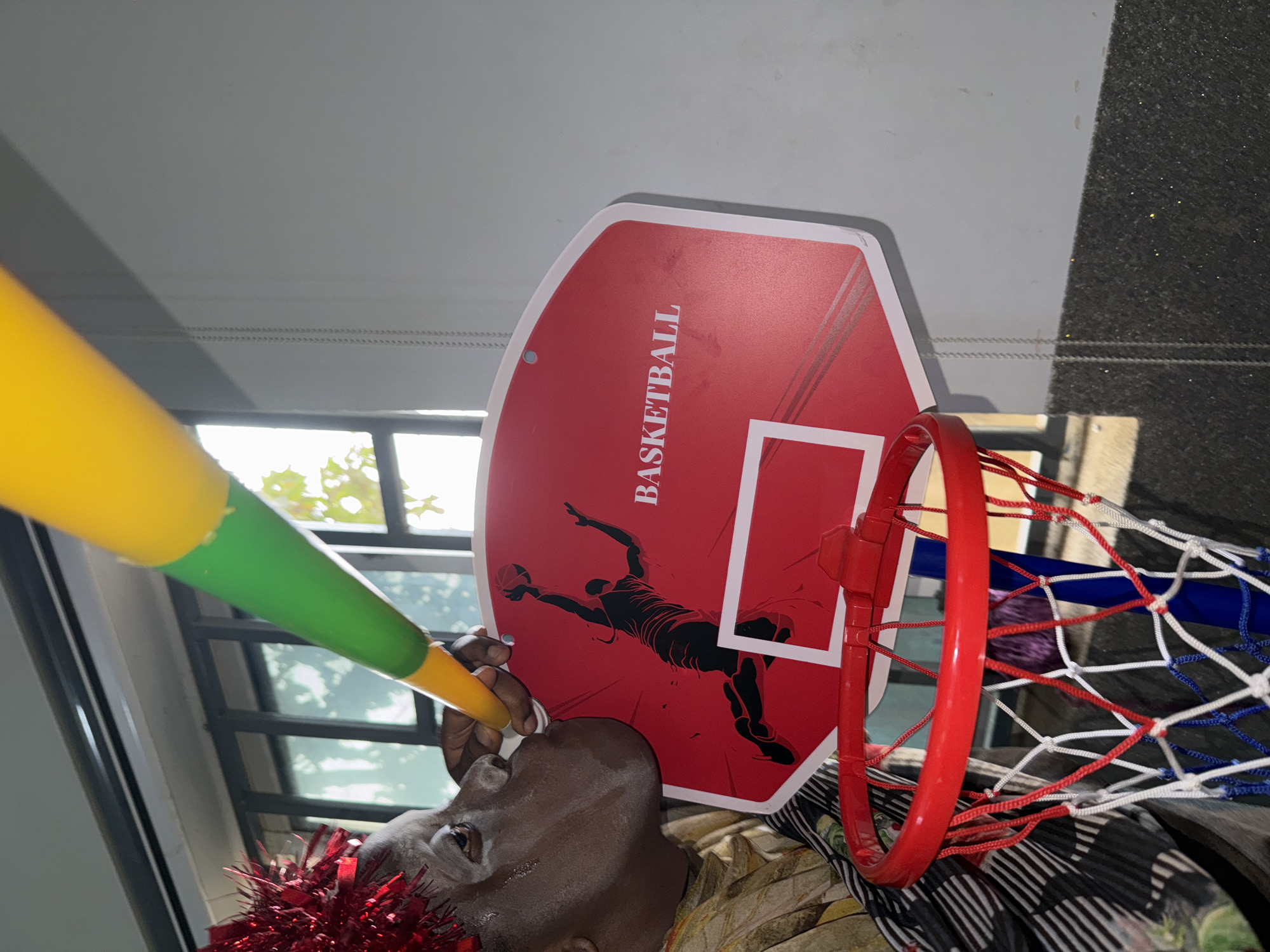 A man is blowing a yellow and green party horn while wearing a red tinsel garland. He is positioned underneath a toy basketball hoop that is decorated with the word "Basketball" and an image of a basketball player. The scene appears to be indoors, possibly near a window, based on the natural light and the presence of window bars. The location is assumed to be in Sabon Tasha, Nigeria.