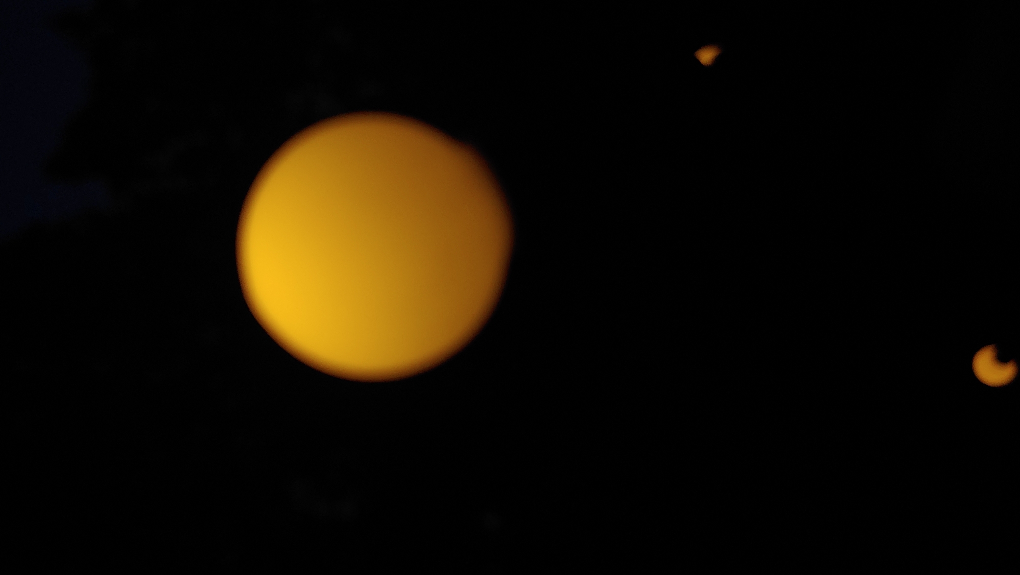 The image is an out-of-focus photograph of a night sky. A large, blurry, orange-yellow disc, possibly the moon or a celestial event like an eclipse, dominates the upper center of the frame. Two smaller, similarly blurry orange-yellow shapes are visible in the lower right and lower left quadrants. The background is almost entirely black, with a hint of dark foliage or a silhouette of trees at the very top edge. There are no people, buildings, or other discernible features of the city of Mukachevo, Ukraine, visible in the image. The focus is entirely on the luminous, abstract shapes against the dark void.