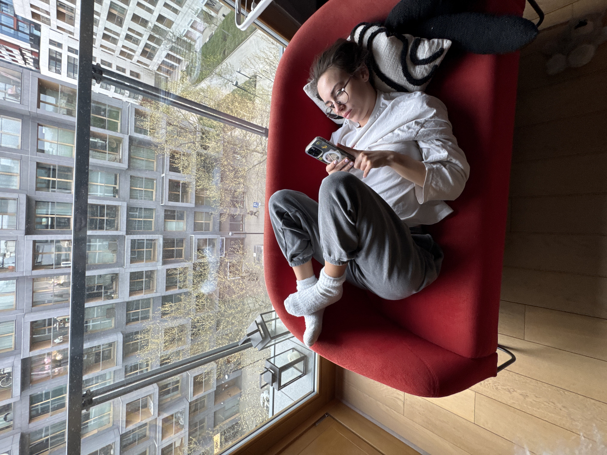 An overhead, slightly tilted shot captures a young woman relaxing on a vibrant red armchair indoors, positioned beside a large window overlooking an urban landscape in Warsaw, Poland. The woman, appearing to be in her 20s or 30s, wears glasses, a white long-sleeved shirt, grey sweatpants, and white socks. She is lying on her left side, propped up by a striped black and white pillow and a dark pillow/blanket, and is intently looking at her smartphone, which she holds with both hands. The phone case has small, indistinct stickers on its back.
Outside the large window, a modern building with a repeating pattern of square-framed windows dominates the view. Trees with delicate, early spring foliage (bare branches with some light green leaves) are visible, suggesting the time of year. Reflections on the window glass show parts of another distant building and the sky, creating a layered visual effect. A prominent dark vertical bar and a horizontal bar intersect the outdoor scenery, likely part of the window's frame or an exterior architectural feature. The indoor space features a light-colored wooden floor.
The scene portrays a quiet moment of leisure during the daytime, indicated by the ample natural light. The woman appears calm and focused on her activity, enjoying a comfortable, private setting against the backdrop of a bustling city.