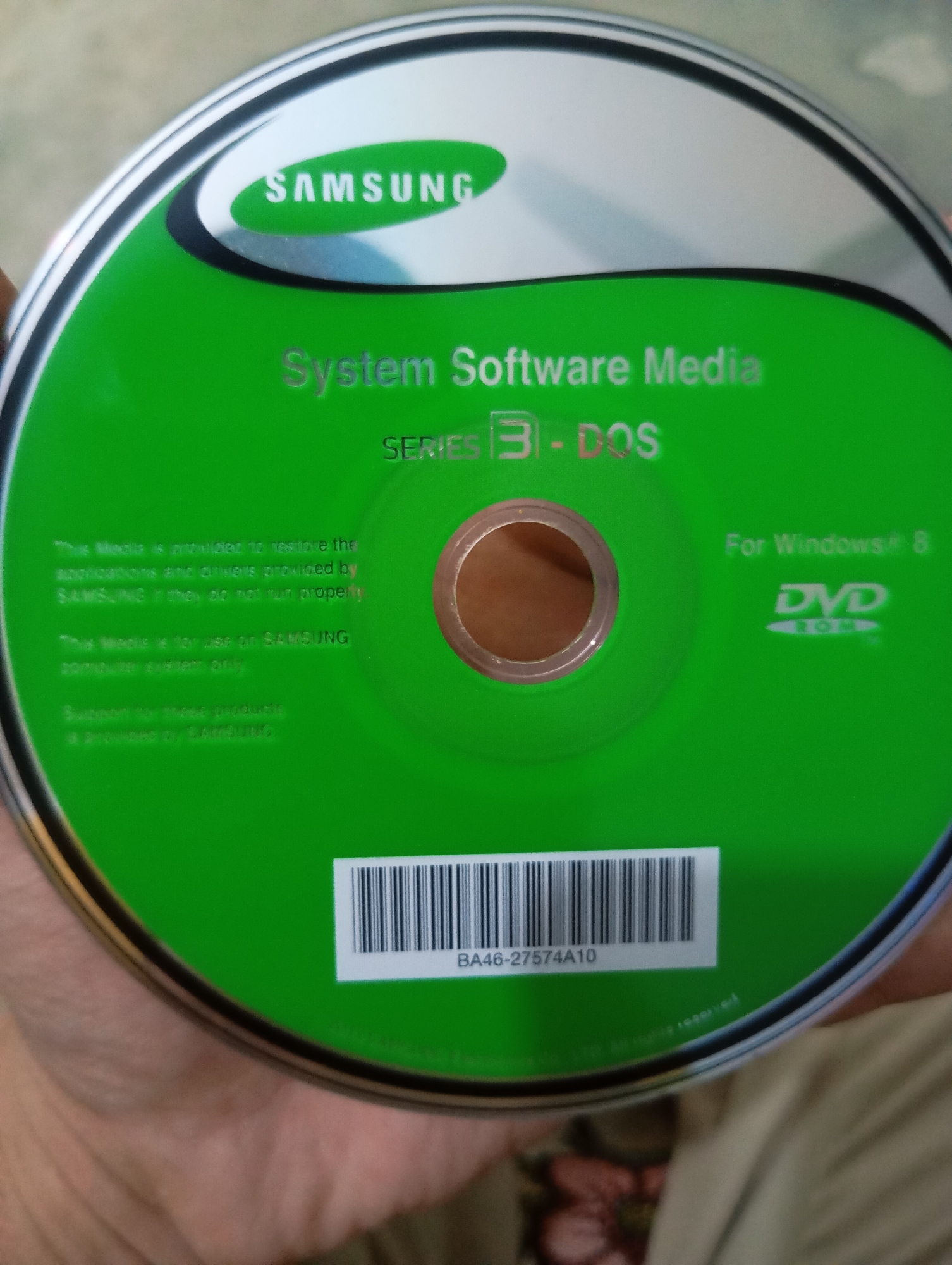 The image shows a DVD disc from Samsung. The disc is green and features the Samsung logo in white. Text on the disc reads "System Software Media" and "SERIES 3 - DOS". There is also text indicating "For Windows 8" and a DVD-ROM logo. A barcode and a serial number "BA46-27574A10" are visible at the bottom. The disc is being held by a person, but only their hand is visible. The background is blurred and out of focus. There are no discernible elements that suggest a specific city or location.