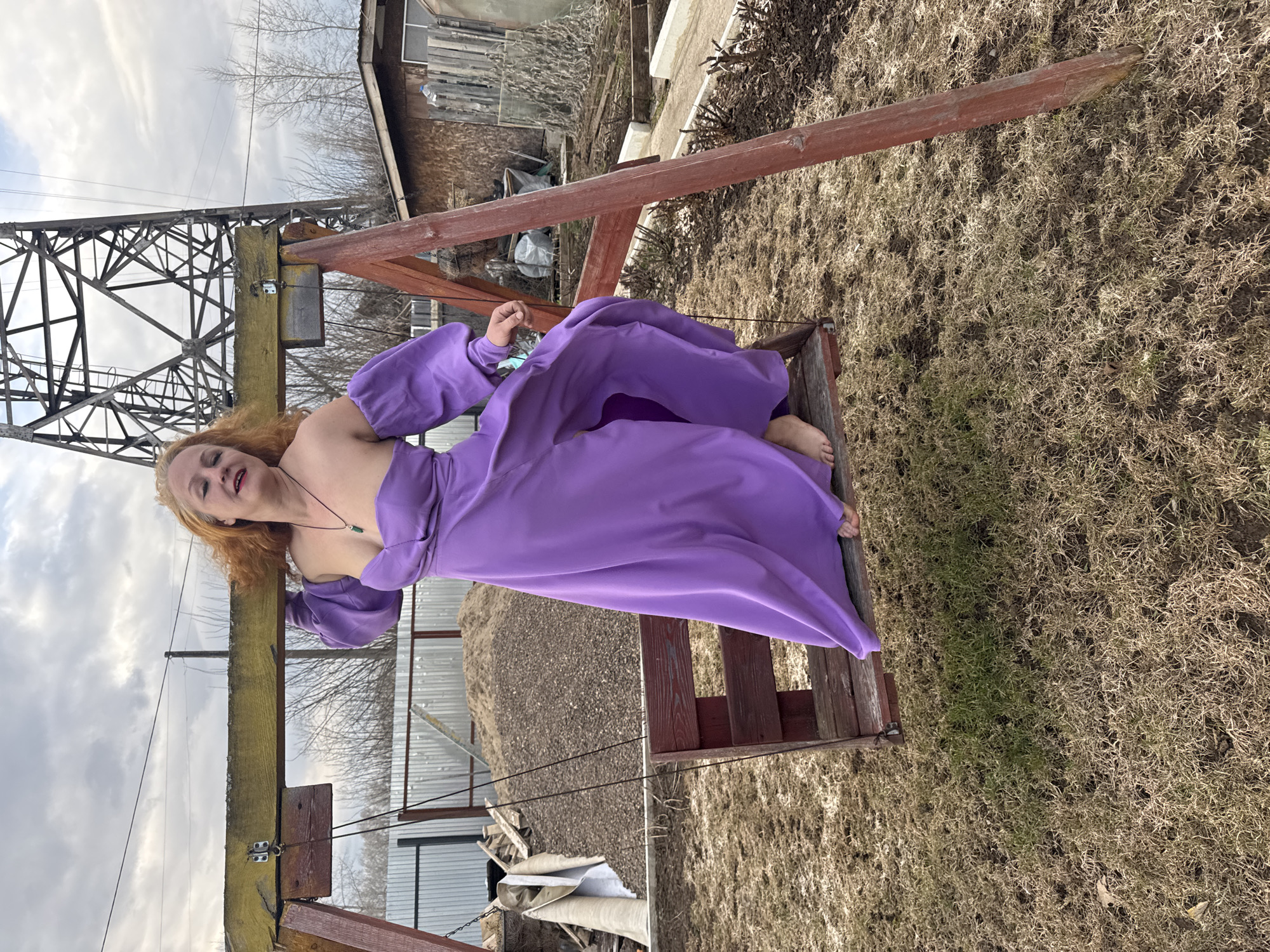 A woman with reddish-blonde hair, wearing a flowing, off-the-shoulder purple dress with puffed sleeves, is seated on a rustic wooden swing. Her bare feet rest on the swing's seat, and she wears a necklace with a dark pendant. She looks towards the viewer with a relaxed expression.
The setting is outdoors, likely a rural or semi-rural area in Chaikovsky, Russia. The ground is covered in dry, yellowish-brown grass, suggesting late winter or early spring. The swing set is constructed from sturdy wooden beams. In the background, a large metal lattice structure, resembling an electricity transmission tower, is prominently visible. Behind the swing, there are several simple wooden buildings or sheds, one with a corrugated metal roof, and bare trees under an overcast, cloudy sky. The time of day is daytime, but the weather appears dull and cloudy. No text is visible in the image.