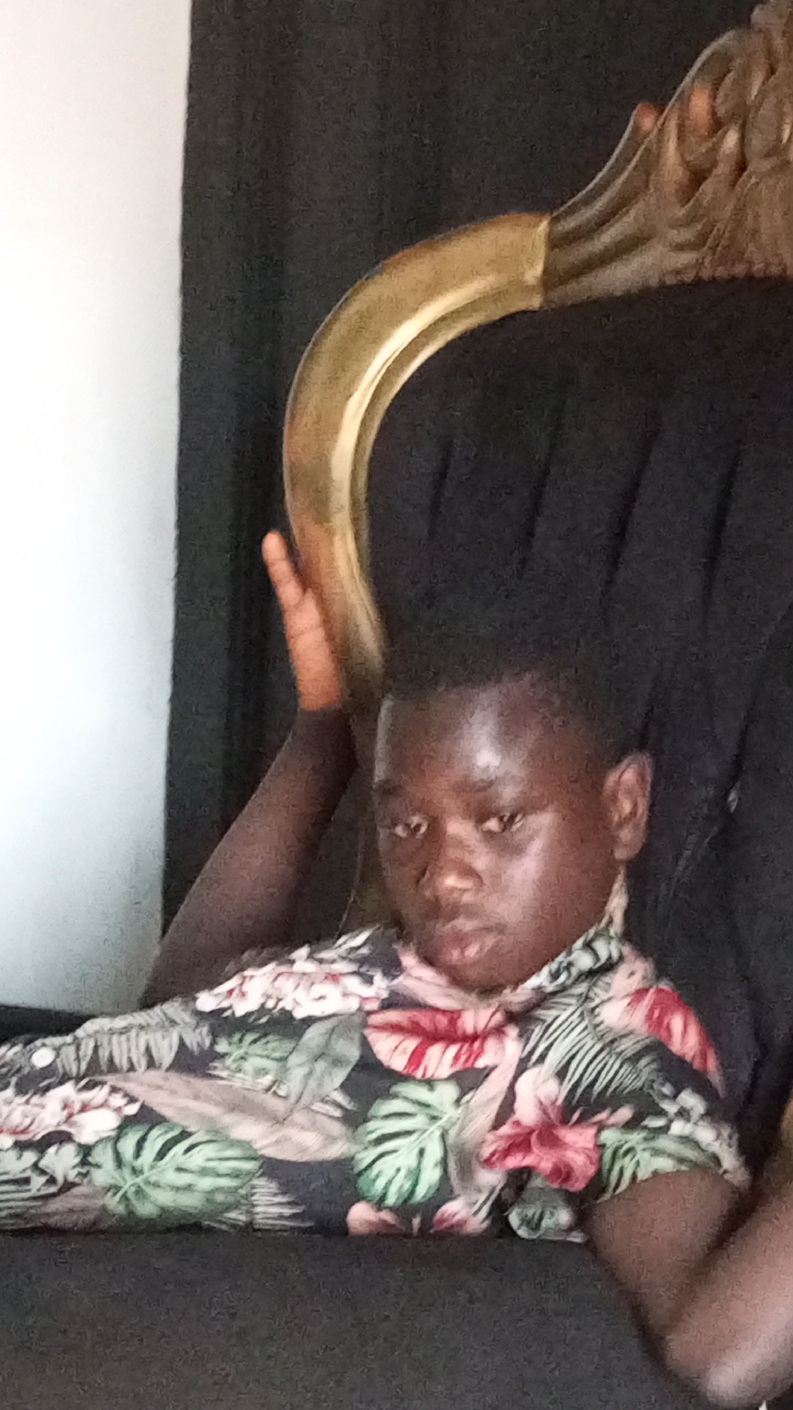 The image features a young boy, likely in Sabon Tasha, Nigeria, lounging on a dark chair with an ornate golden frame. The boy has dark skin and short, dark hair. He is wearing a dark-colored shirt adorned with a vibrant floral pattern, including green leaves and red and pink flowers. His expression is neutral to slightly serious as he looks directly at the viewer. One of his arms is bent, with his hand resting near the golden, curved armrest of the chair. The background shows a dark curtain on the right side of the image and a plain white wall on the left, indicating an indoor setting. The lighting suggests ambient indoor conditions. There is no visible text in the image.