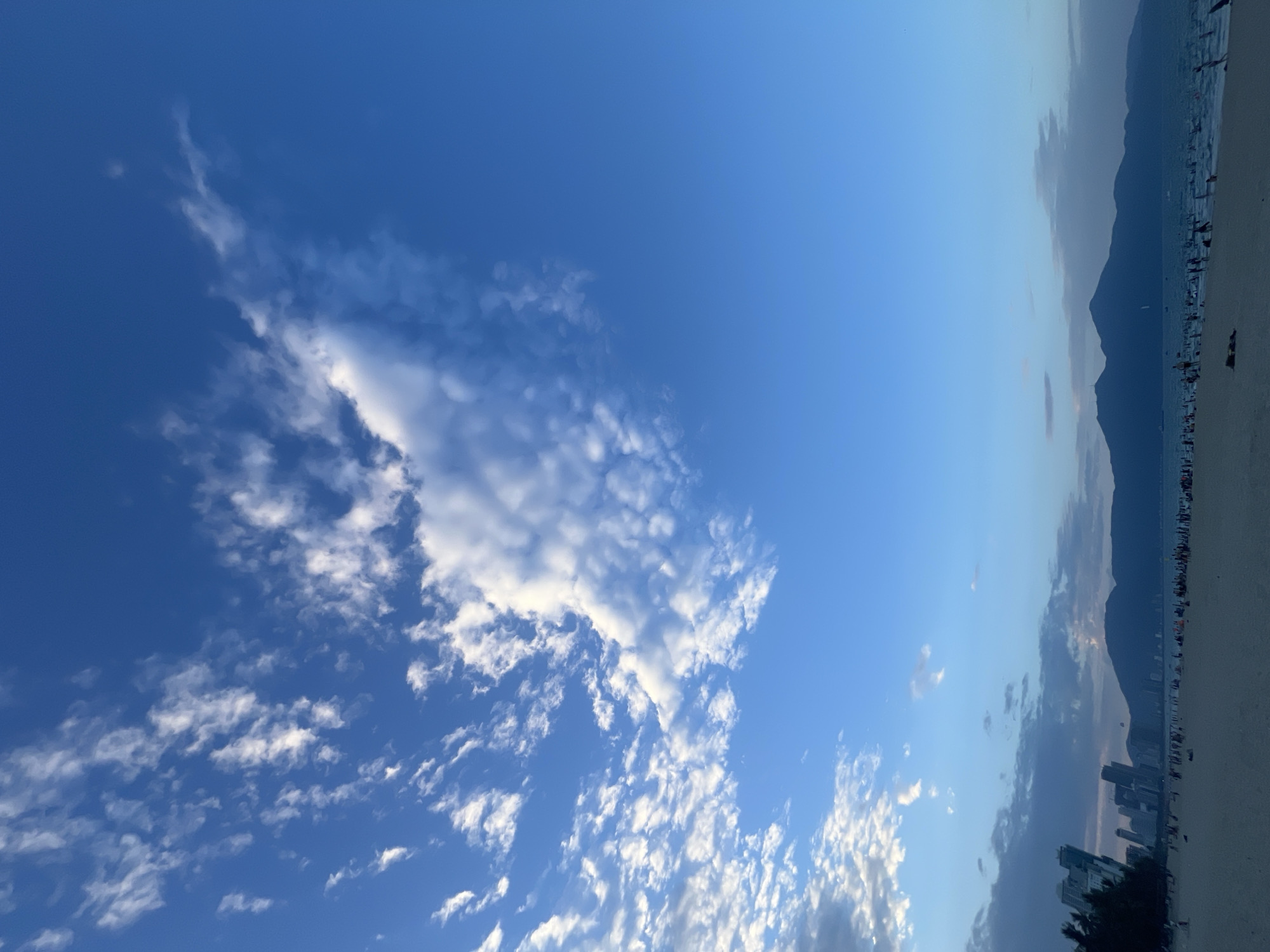This image depicts a wide, sandy beach in Da Nang, Vietnam, with a vibrant blue sky overhead scattered with white, wispy clouds. In the distance, a silhouette of a city skyline with tall buildings is visible on the left, transitioning to a range of dark, silhouetted mountains on the right. The ocean stretches out beyond the beach, and numerous people are scattered across the sand and in the shallow water, engaged in various leisurely activities. The overall atmosphere suggests a serene beach scene, likely in the late afternoon or early evening due to the lighting.