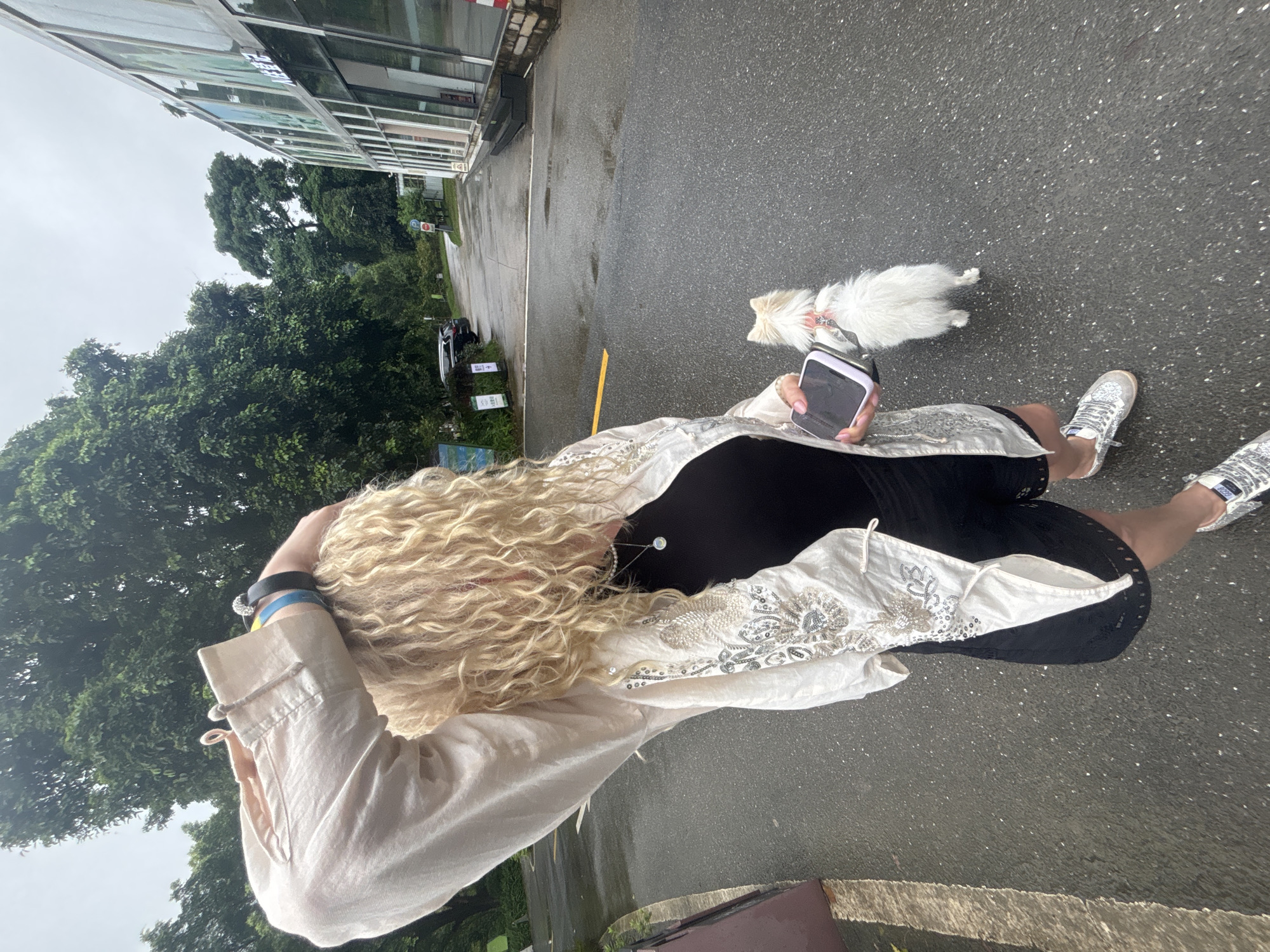 A woman with blonde curly hair stands on a wet asphalt road, holding a smartphone and a small white dog on a leash. She is wearing a black tank top, black shorts with a decorative hem, a light-colored open jacket with intricate silver embroidery, and silver sneakers. Her left arm is raised, touching her head, and she has several bracelets on her wrist. The dog is facing away from the camera. In the background, there are lush green trees, a modern building with large glass windows, and parked cars on the side of the road. The sky is overcast. The scene appears to be outdoors, possibly in a park or near a public facility. The location is identified as Guangzhou City, China.