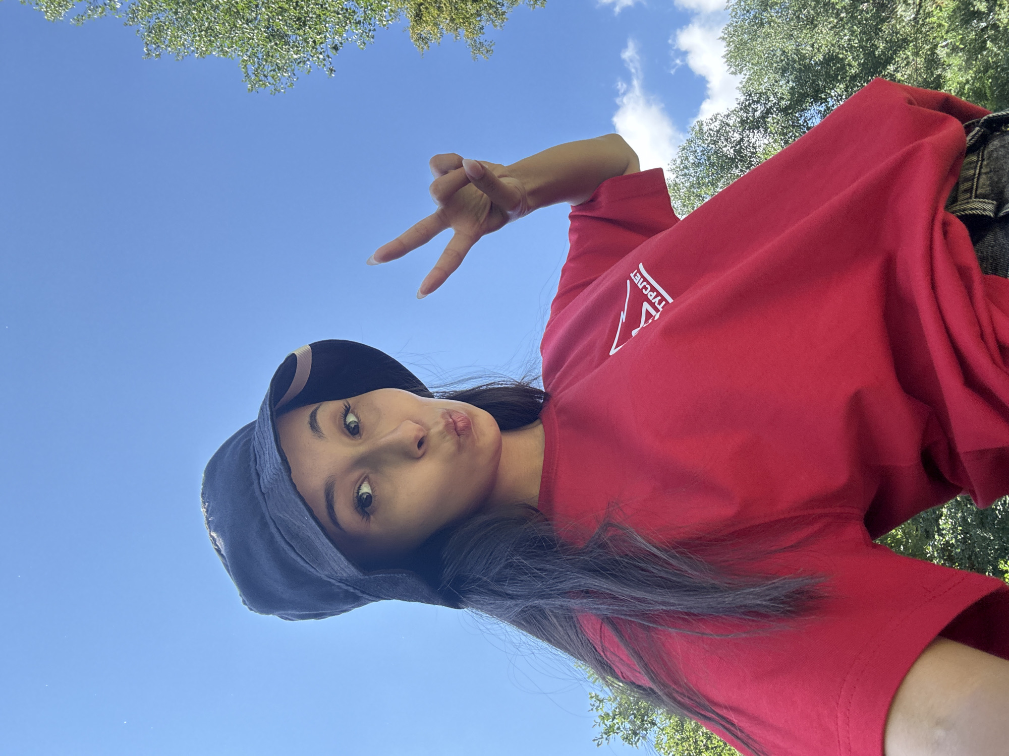 A young woman wearing a red t-shirt and a dark bucket hat takes a selfie outdoors. She is making a duck face and holding up two fingers in a peace sign. The background consists of a clear blue sky with a few scattered clouds and green trees. The location is Doma 1096 Km, Russia.