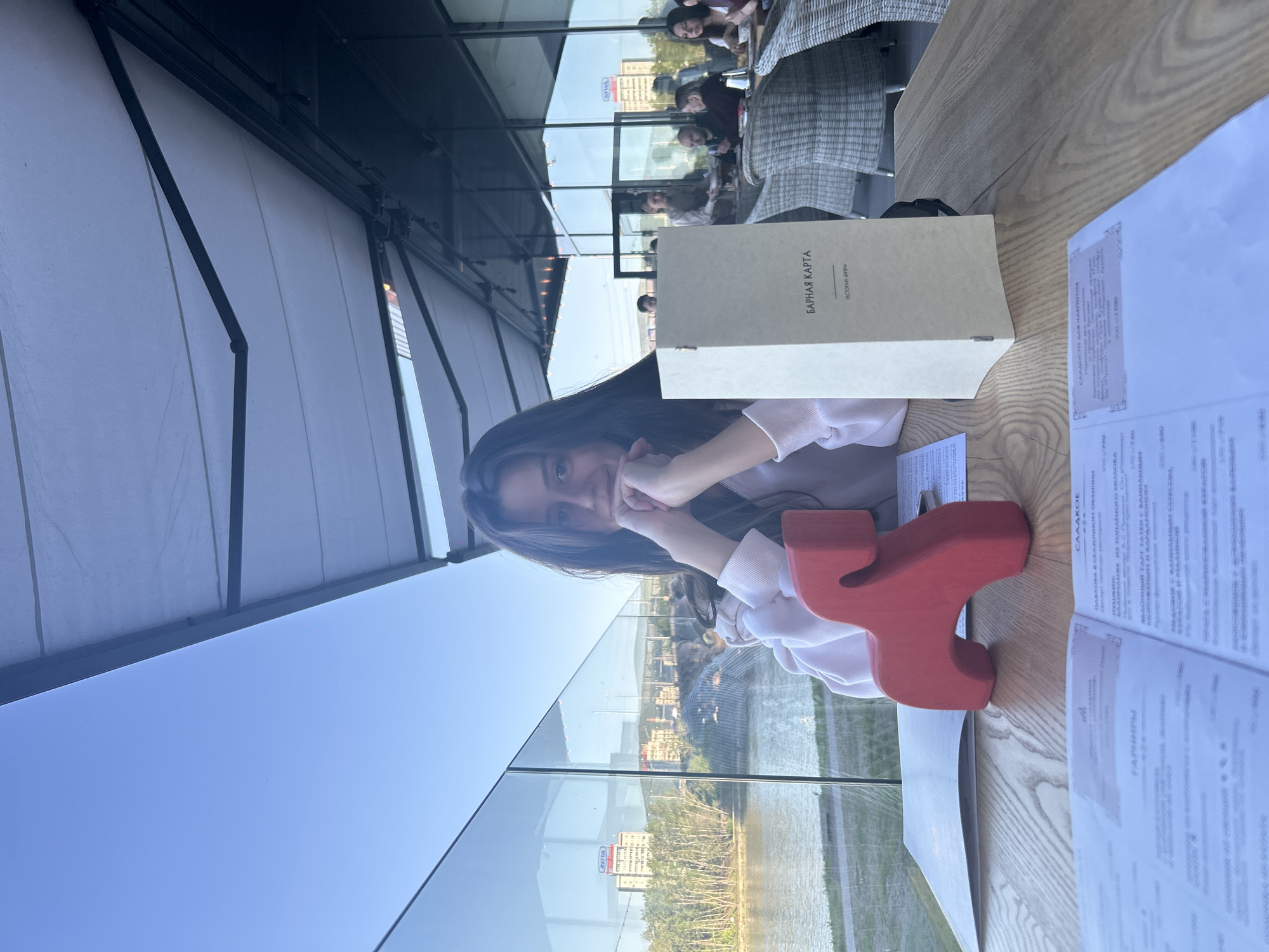 Here is a concise description of the image:
A young woman sits at a table on an outdoor patio in Krasnoyarsk, Russia, in a restaurant called "Буян". She rests her face in her hands, appearing thoughtful. On the table are a menu, a drinks list, a red decorative object, and what may be a city view beyond a glass barrier. Other patrons are visible at tables in the background.