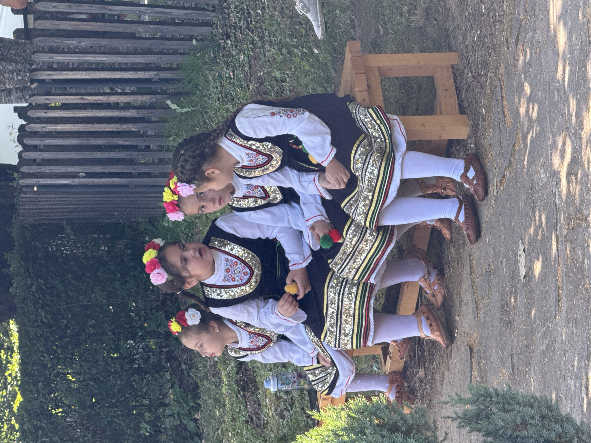 Four young girls, dressed in traditional Bulgarian folk costumes, are seated on a wooden bench outdoors. They are positioned side-by-side, facing the viewer's right. Each girl wears a white blouse with embroidered cuffs, a dark vest adorned with sequins and embroidery around the neckline and hem, and a skirt with striped trim and sequined detailing. They also wear white tights and brown leather shoes. Flower crowns are on their heads. The girl second from the left is holding a yellow pom-pom, and the girl on the far right is holding a green and red pom-pom. A blue water bottle with a cartoon character is visible on the ground to the left of the bench. The background consists of lush green bushes, a wooden fence, and trees. The ground is a mix of dirt and gravel. The image appears to be taken in Ribaritsa, Bulgaria, likely during a cultural event or performance.