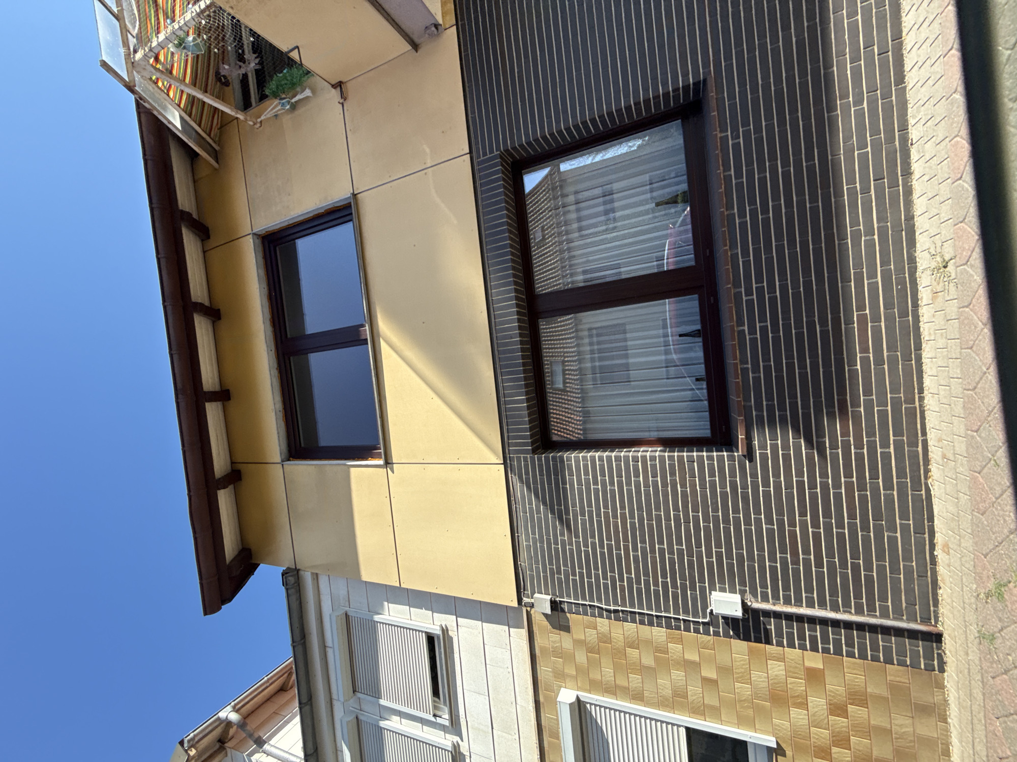 The image shows the exterior of a multi-story building on a sunny day. The building has a light yellow facade on the upper stories and dark grey brick on the lower story. There are several windows visible, some with closed white roller shutters and others with sheer white curtains. The windows have dark brown frames. A small awning with a colorful striped fabric is attached to the upper right side of the building. A metal pipe runs vertically along the dark brick wall, connected to a small white electrical box. The ground in front of the building is paved with interlocking bricks. The sky is clear and blue. This scene is located in the city of Bad Sachsa, Germany.