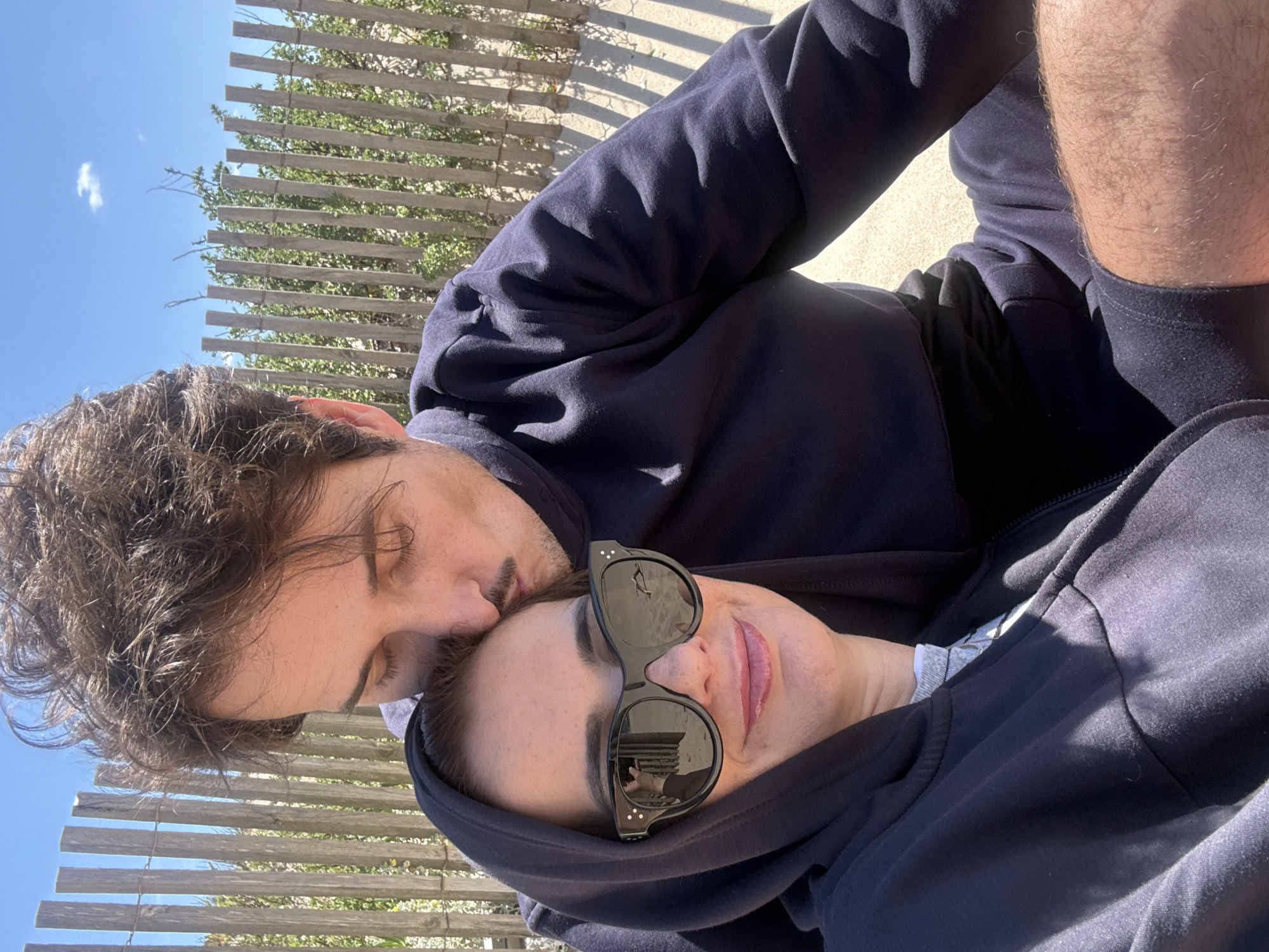 The image shows two people in close proximity, likely a couple. One person is kissing the other on the forehead. The person being kissed is wearing sunglasses and a dark hoodie. Both are wearing dark clothing. The background features a wooden fence and glimpses of a clear blue sky with a small cloud, suggesting an outdoor location. The beach in Vila Nova de Gaia, Portugal.