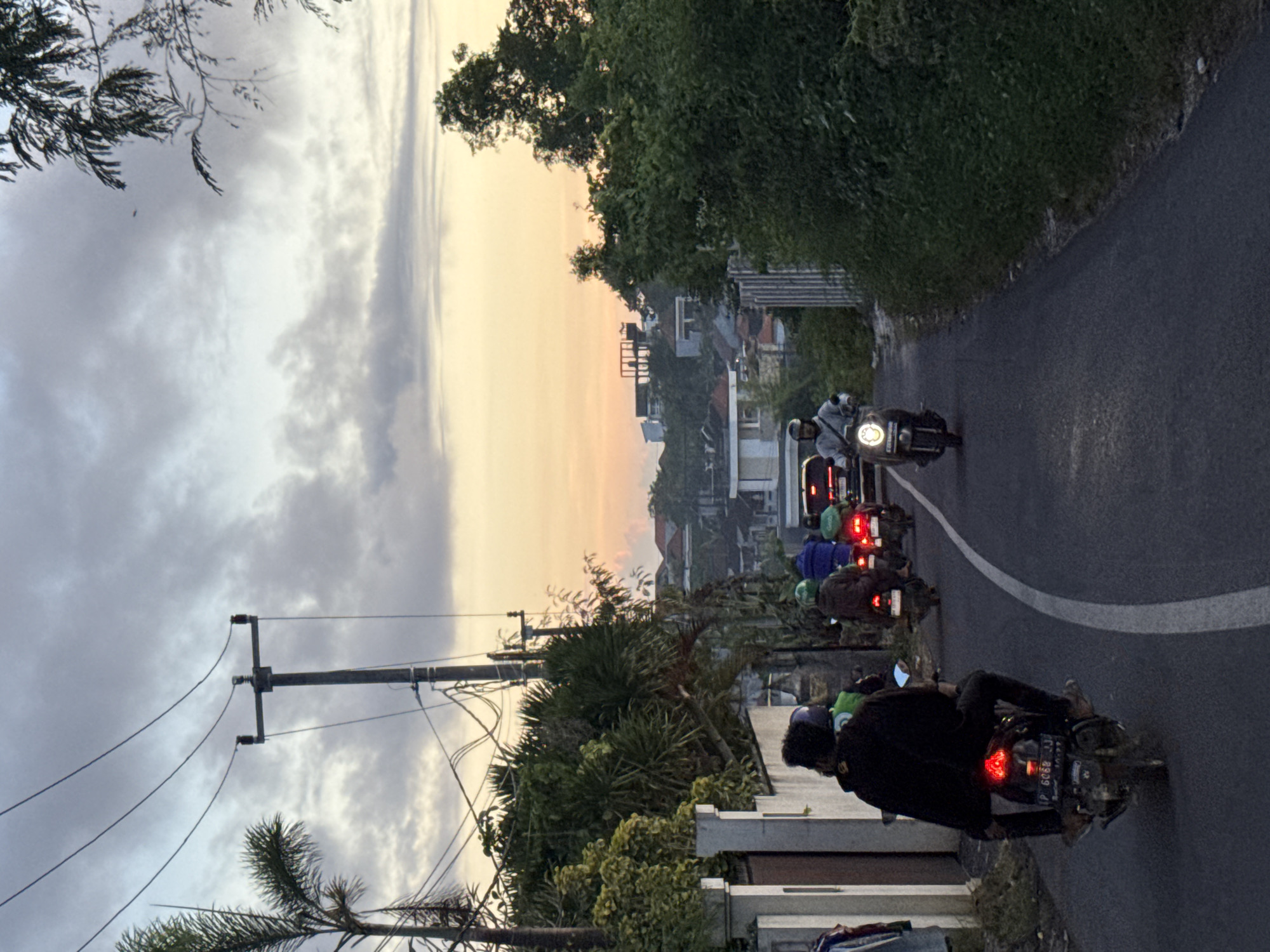 The image depicts a road scene at what appears to be dusk or dawn, given the warm, orange-yellow glow on the horizon transitioning to a cloudy, grey sky overhead. The setting is likely a residential or semi-urban area in Kuta Selatan, Indonesia, judging by the architecture, dense tropical vegetation, and prominent use of motorbikes.
Several individuals are visible on motorbikes, either riding or stopped in traffic on the asphalt road. In the foreground, a person wearing a dark jacket and helmet is seated on a motorbike, its red tail light illuminated. The motorbike's license plate, "DK 6088 AJ," is clearly visible, with "DK" being the vehicle code for Bali, consistent with the Kuta Selatan context. Further down the road, another person in a white top and helmet rides a motorbike with its headlights on, while a line of other motorbikes, some with their brake lights illuminated, follows behind. Some riders wear vests or helmets with green accents; one appears to have "GRAB" branding, indicating a ride-hailing service.
The road features a white curved line marking a lane or edge. On either side, lush green foliage and trees line the path, with residential buildings visible in the background, some featuring red-tiled roofs. A prominent utility pole with multiple power lines extends across the left side of the frame, contrasting against the cloudy sky. The overall impression is one of everyday travel or commuting in a busy, tropical locale during a transitional period of the day.