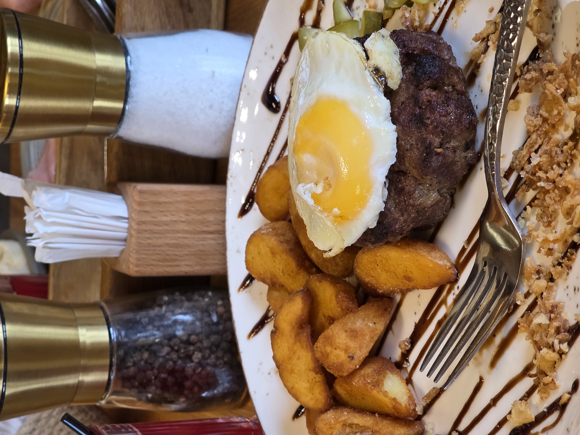 A top-down, close-up shot showcases a plate of food, likely a hearty meal, set against a wooden surface. The meal consists of a thick, grilled meat patty topped with a sunny-side-up egg, its golden yolk glistening. Accompanying the patty are a generous portion of golden-brown potato wedges, some dusted with salt. Drizzled across the plate are thin lines of a dark sauce, possibly balsamic glaze or a reduction, adding a touch of elegance. Scattered around are crispy fried onions, providing a textural contrast. A metal fork rests on the right side of the plate, its tines poised to dig in.
To the left of the plate, out of focus, are two glass grinders with gold accents, one filled with white salt and the other with dark peppercorns. Next to them, a wooden holder contains a stack of white paper napkins. The overall impression is of a rustic yet appealing meal, possibly served in a restaurant or a home kitchen. The lighting suggests daytime, and the focus is entirely on the food, implying a moment captured during a meal. There is no visible text in the image. The setting appears to be indoors, with a warm, inviting atmosphere suggested by the wooden elements.