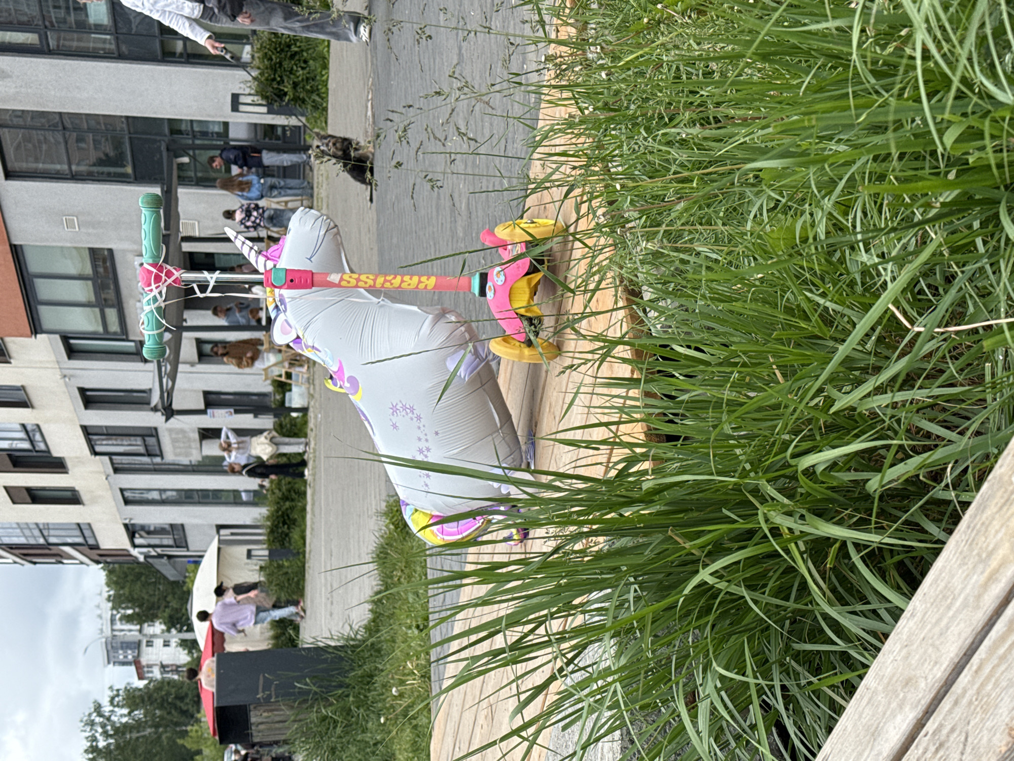 The image depicts an outdoor urban scene in Izhevsk, Russia, on an overcast day. In the foreground, a vibrant green grass patch partially covers a wooden plank surface. Prominently positioned on this wooden surface is a pink and yellow three-wheeled child's scooter, identifiable by the "KRISS" branding on its stem. Attached to the scooter's handlebars is a large, deflated or partially deflated, grey inflatable unicorn balloon, featuring a rainbow-colored horn and tail, and subtle purple star-like patterns on its body.
Behind the wooden platform, a paved pedestrian area extends into the midground. A small, dark-colored dog, possibly a Yorkshire Terrier, is sitting attentively on this paved surface, facing left. To the far right, partially visible, is a person wearing grey pants and a white long-sleeved top, holding a leash connected to the dog.
The background is dominated by modern, multi-story residential buildings with a mix of light and dark facades, characterized by numerous large windows. Several groups of people are scattered across the midground and background:
- On the left side, near white tents and a red umbrella, individuals are gathered, with one person in a light purple shirt walking away.
- Towards the center-right, a group of approximately five people are standing and conversing in front of the buildings, with one individual holding their hands to their head. Another smaller group stands near a building entrance further back.
- Trees and additional buildings are visible in the distant background under a cloudy sky.