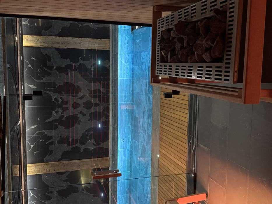 The image captures a modern and luxurious interior, likely a high-end sauna or spa area, consistent with a setting in Courchevel, France. No people are visible, emphasizing the serene and unoccupied nature of the space.
The scene features a sophisticated design blending dark, polished surfaces with natural wood and illuminating elements. A prominent feature is a contemporary sauna heater positioned in the upper right, enclosed in a light wooden frame with a metal grate, and filled with dark sauna stones. Below this, a slatted wooden wall adds warmth and texture to the environment.
The main wall on the left showcases dark, marble-patterned tiles with lighter, flowing veins, creating a dramatic backdrop. Reflected on this surface are thin, vertical reddish lines and subtle light sources, suggesting careful architectural lighting. Clear glass partitions, secured by sleek black hinges, divide the space, offering an open yet defined layout.
A striking blue illuminated vertical column or feature is visible towards the center-right, glowing brightly and hinting at a water feature like a cold plunge pool or an aesthetic light installation, characteristic of a spa. The floor and lower wall sections are covered in dark, solid rectangular tiles, maintaining the elegant and minimalist aesthetic. Subtle embedded lights create reflections and highlight the glossy surfaces throughout the room.
The overall impression is one of modern luxury, impeccable cleanliness, and a tranquil atmosphere designed for relaxation and wellness. No visible text is present in the image.