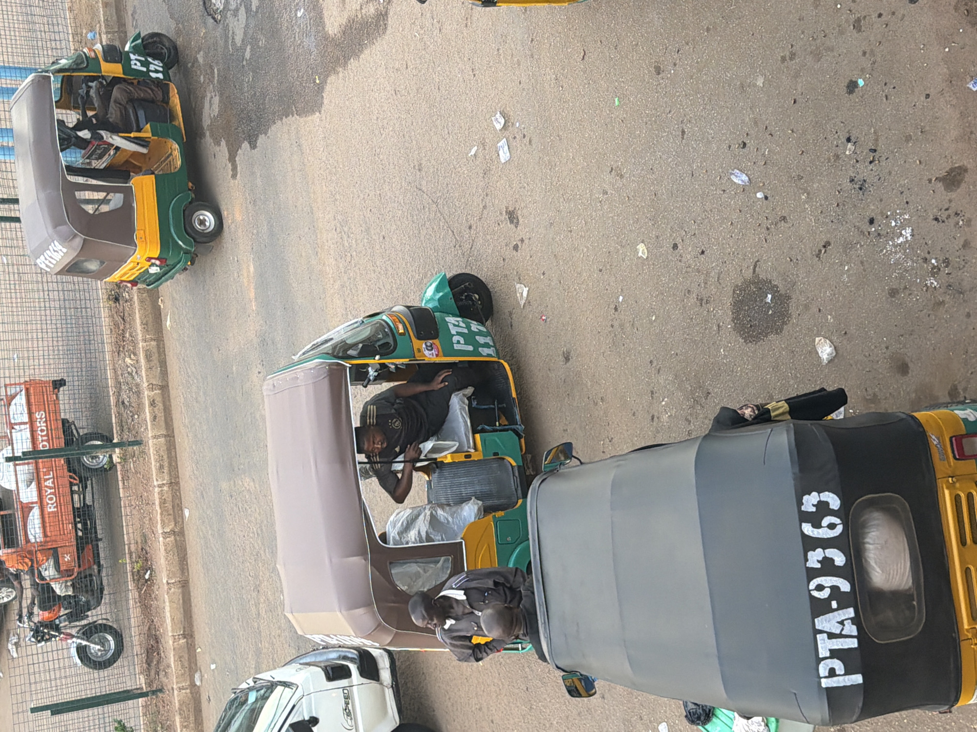 In Bukuru, Nigeria, on a paved street with scattered debris, an overhead view captures three auto-rickshaws. The foremost vehicle, seen from above, is gray and black with "PTA-9363" stenciled in white on its rear. Two men are visible within this rickshaw, one seated and the other standing. Adjacent to this, a green, yellow, and brown auto-rickshaw is partially visible, with "PTA 117" on its front fender and a driver in a dark t-shirt. Further in the background, a separate orange auto-rickshaw with a flatbed is parked. This vehicle has "ROYAL MOTORS" written on its side. The scene suggests a commercial area with active transportation.
