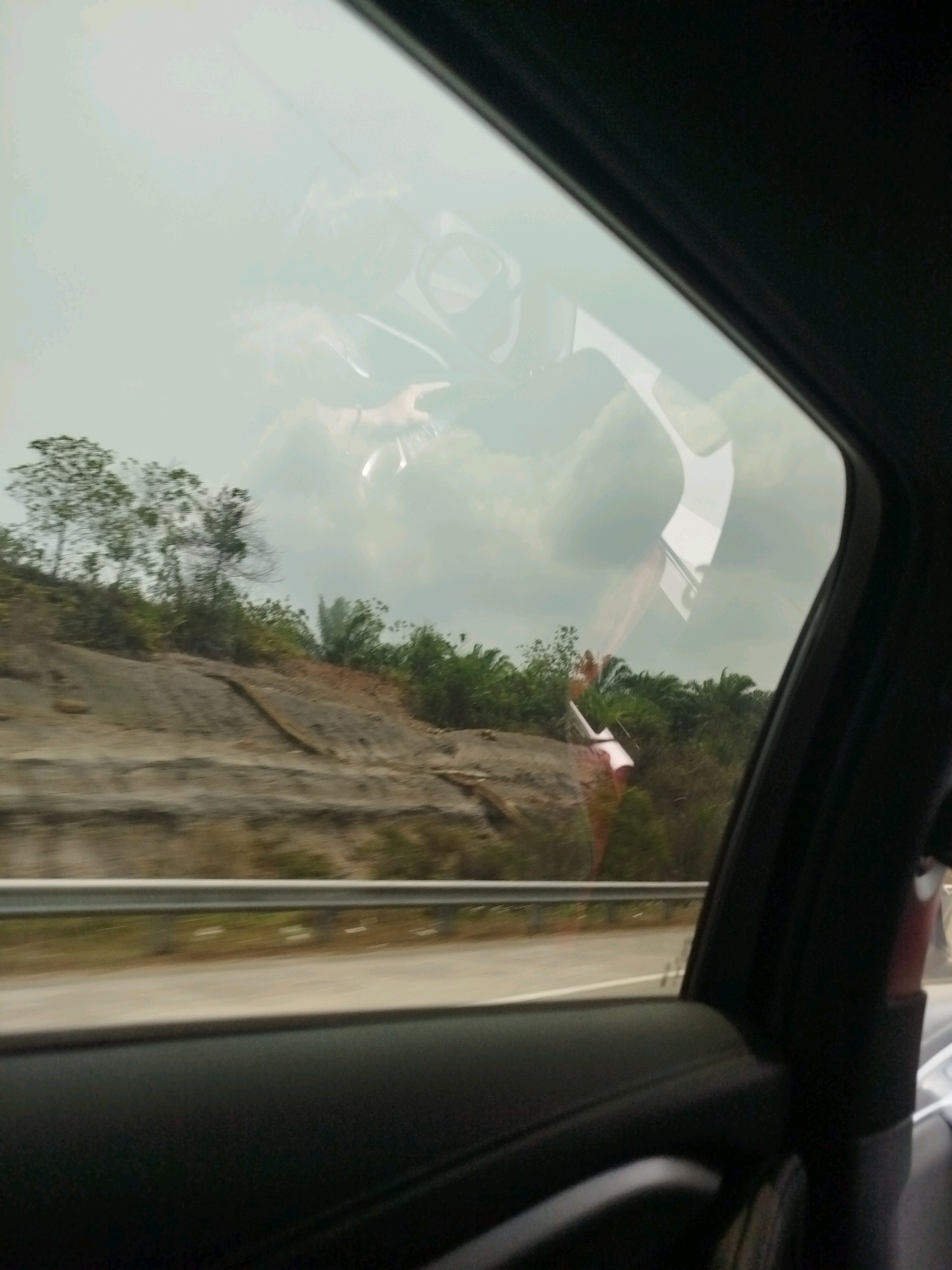 The image shows a reflection on a car window, obscuring the view of the outside landscape, which appears to be a rural area with hills, vegetation, and a road. The reflection shows the interior of the car, including what seems to be a person or people, and possibly parts of the car's dashboard and seats. The lighting suggests it is daytime. The provided crops highlight different areas of the reflection and the partially visible exterior. There is no specific information that can identify the location as Bukit Merdeka, Indonesia.