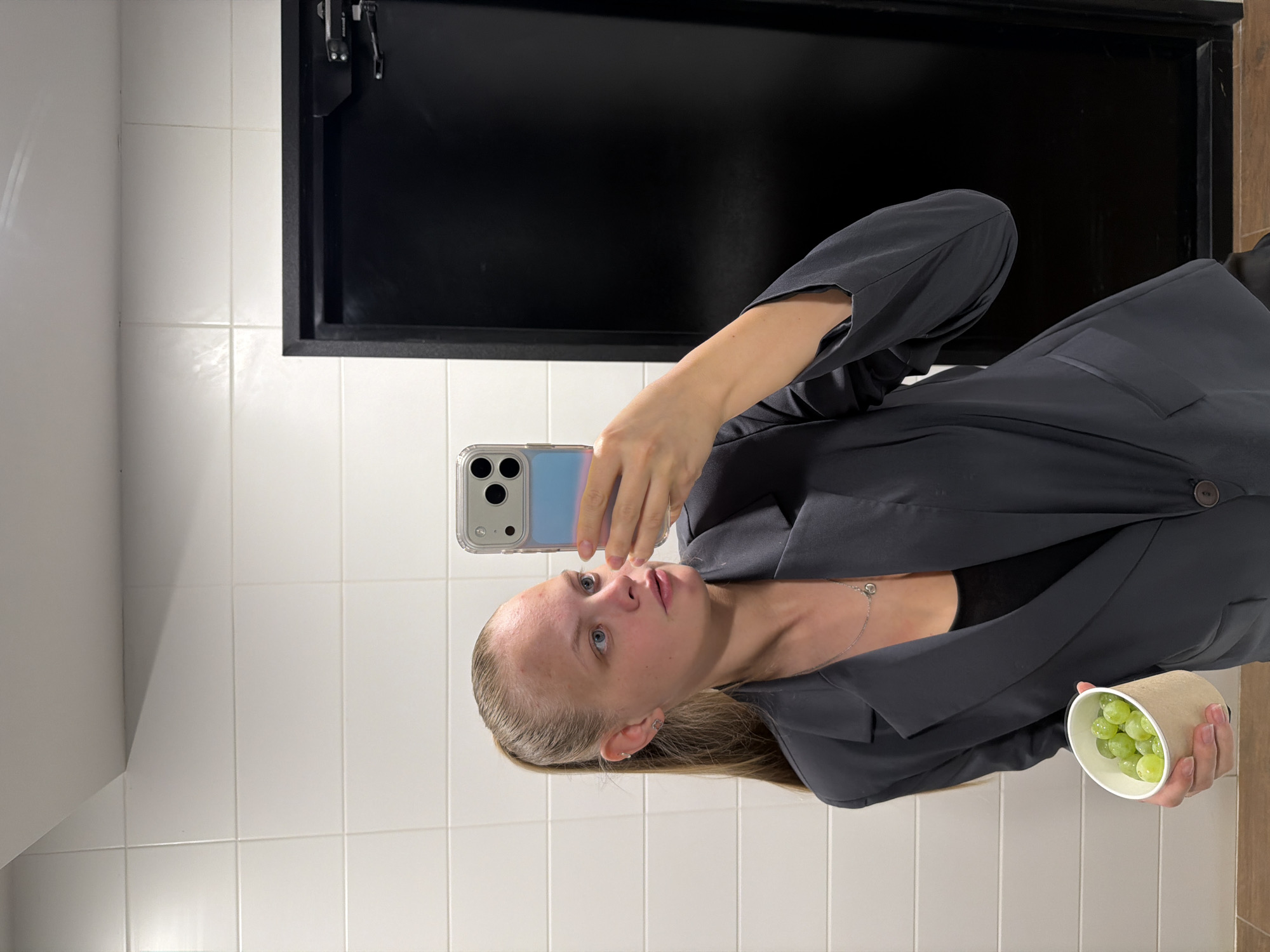 A woman is taking a selfie in what appears to be a bathroom. The background consists of white tiled walls and a black rectangular element resembling a mirror or panel with a metallic hinge. The woman is wearing a gray blazer over a black top, and she is holding a container with green grapes in her right hand. She is holding a smartphone in her left hand to take the picture. There is no indication of this being in Izhevsk, Russia.