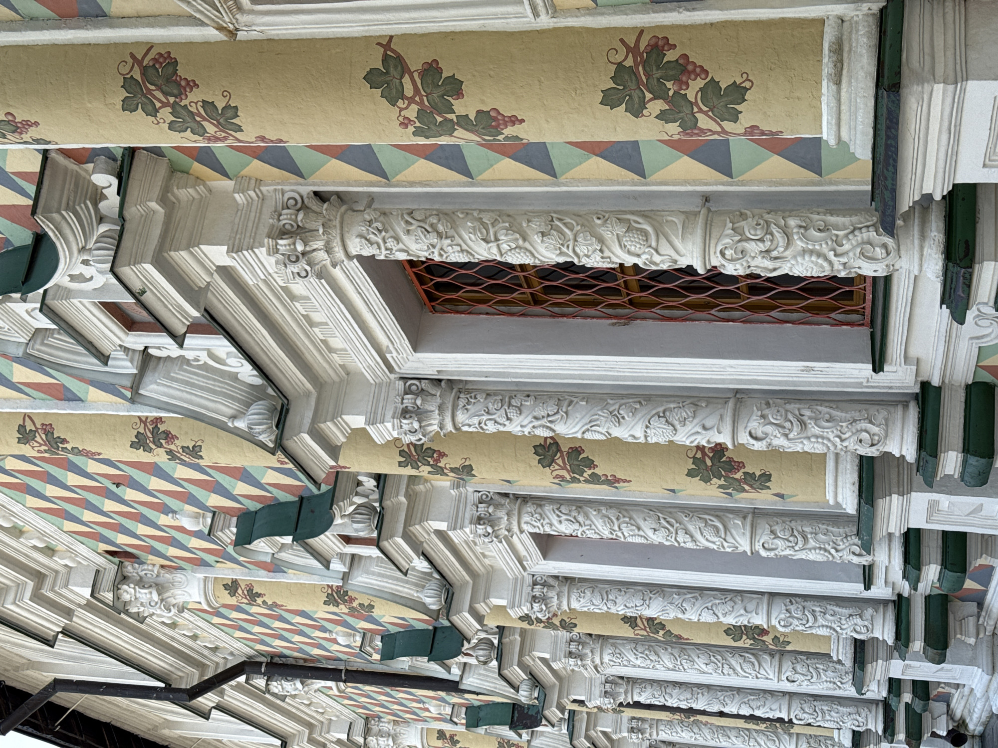 The image showcases a highly ornate, angled ceiling with repeating patterns and decorations. The ceiling features alternating bands of yellowish-tan with grape and vine motifs and geometric, triangular patterns in green, blue, yellow, and red. Detailed white plasterwork, including molding and intricate designs, adorns the edges and structural components. One section includes a window covered with an orange-red mesh screen. Green accents are visible on some of the supporting structures. The image is taken from a low-angle perspective, looking up at the ceiling. Location: Unknown, Unknown.