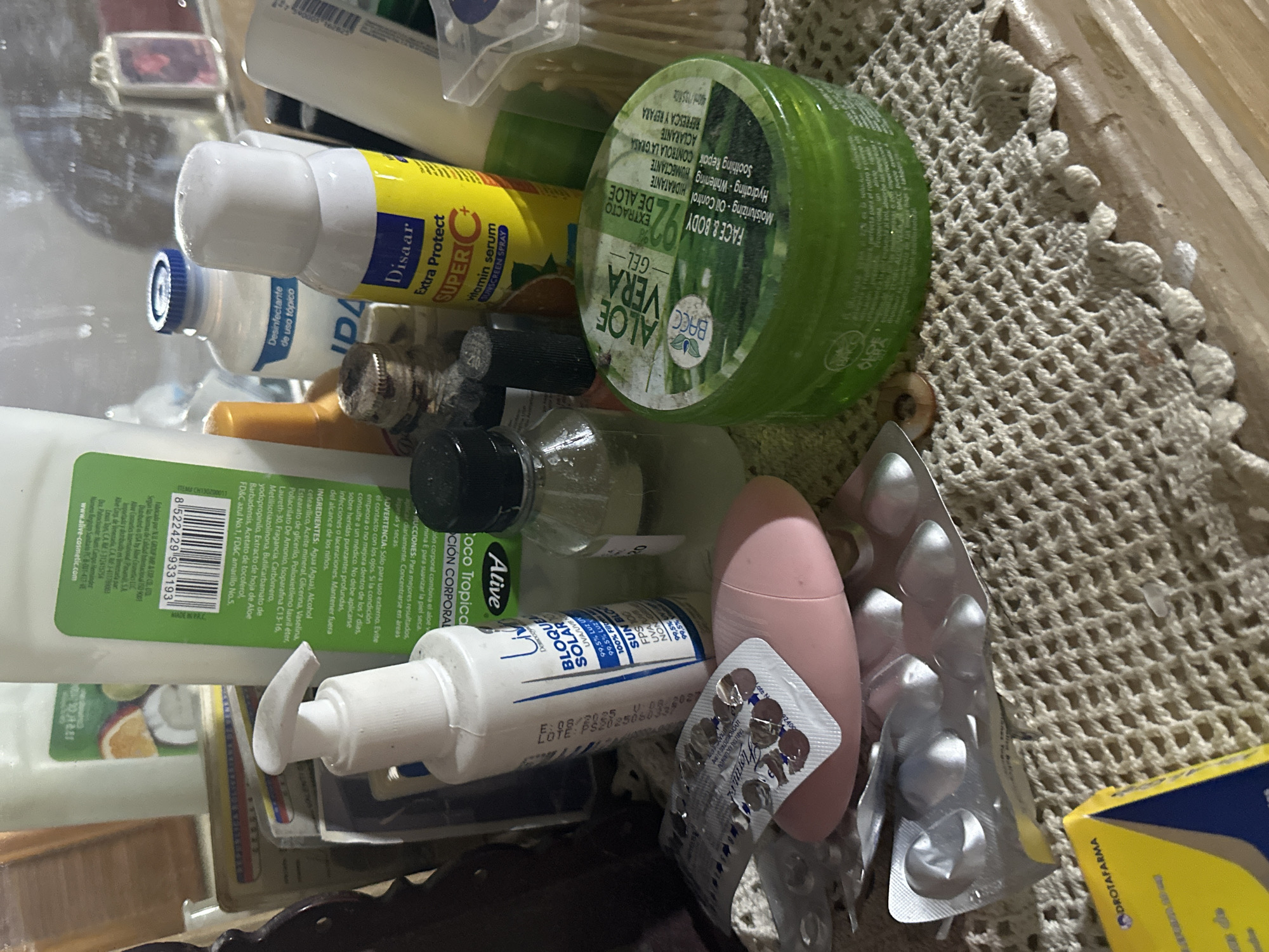 This image depicts a collection of personal care products and medications situated on a light-colored, crocheted lace mat, which rests upon a light-brown wooden surface, likely a dresser or vanity. The scene is presented as a domestic setting, potentially located in the city of Juan Griego, Venezuela.
In the foreground, a yellow and blue cardboard box, partially visible on the bottom left, displays the text "DROTAFARMA." Adjacent to it, a white pump-dispenser bottle, branded "Liv Dermocosméticos," is labeled as "BLOQUE SOLAR" (sunscreen) with visible claims of UVA/UVB and visible light protection, and "100% FILTRO SUN BLOCK." To the right of this bottle, two blister packs are present: one partially used, containing several round brown pills in some compartments and others empty, and another, mostly empty, crumpled silver foil blister pack. A smooth, pink, oval-shaped object is positioned among these items.
Further to the right, a small, clear glass bottle with a black screw-top cap contains a transparent liquid. Beside it stands a large, circular, green container of "BACC ALOE VERA GEL," labeled as containing "92% EXTRACTO DE ALOE" and marketed for "FACE & BODY," with listed benefits including moisturizing, oil control, hydrating, whitening, soothing, and repairing. To the right, a yellow spray bottle is labeled "Disaar Extra Protect SUPER C vitamin serum Sunscreen Spray." Immediately next to it is a white bottle with a blue cap, bearing the label "IPA Desinfectante de uso tópico" (topical disinfectant). On the far right, a clear plastic container holds a quantity of white cotton swabs.
In the background, a tall, white pump-dispenser bottle features a green label with a visible barcode (8522429933193) and text indicating "Coco Tropical" and "Ative," suggesting it is a body lotion. Behind these objects, a reflective surface, possibly a mirror, shows blurred reflections, including what appears to be a small framed picture or decorative charm.