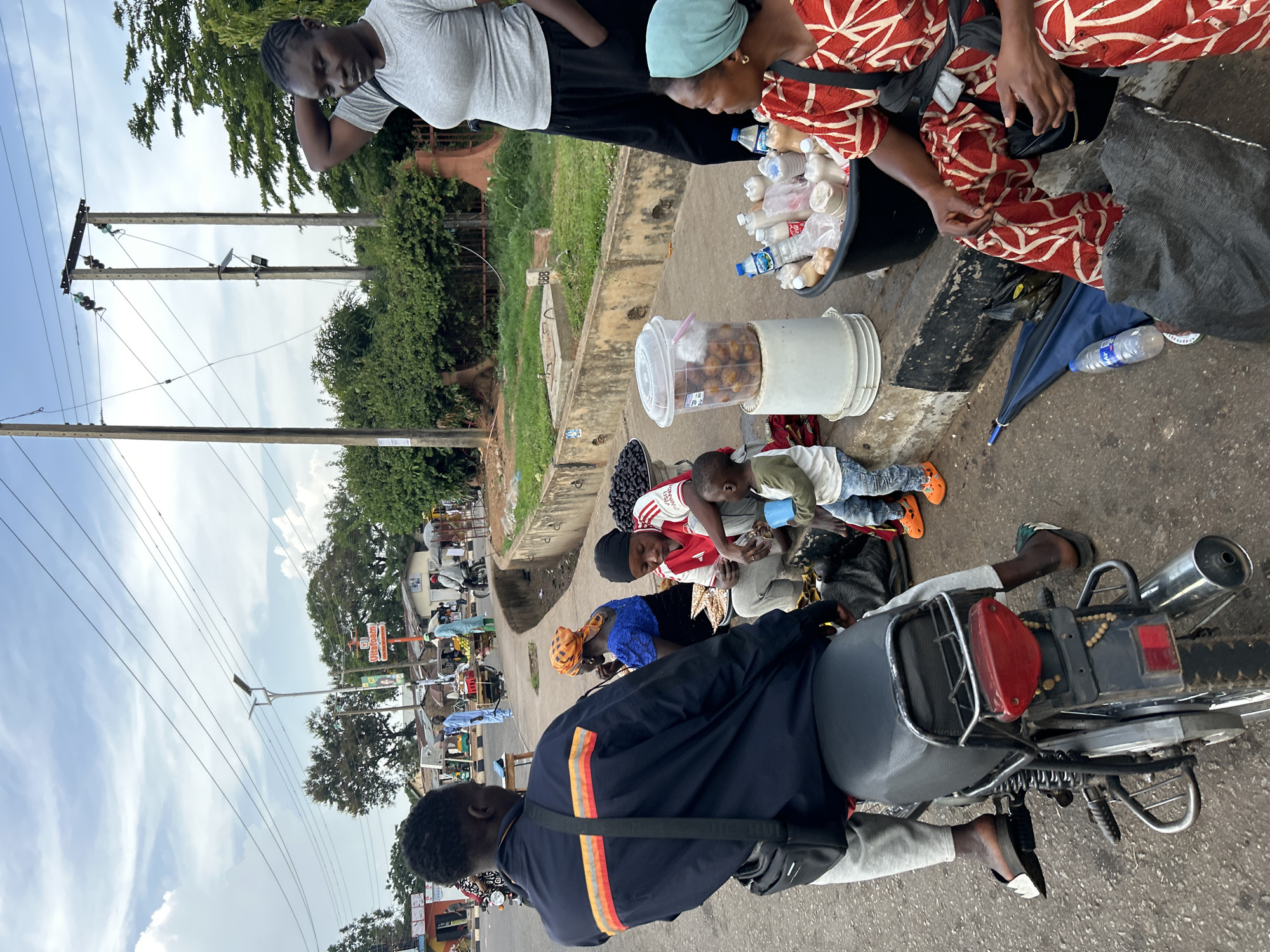 The image depicts a street scene in Jos, Nigeria, with several individuals engaged in commerce and daily life. In the foreground, a person is seated on a motorcycle. To their right, two women are selling goods, including beverages in bottles and what appear to be dried fruits or nuts. A young child is also present, holding a blue cup. Behind them, other people are visible, some tending to market stalls with produce. The environment includes a roadside with a curb, a paved sidewalk, and overhead power lines, suggesting an urban setting. The overall atmosphere appears to be that of a bustling marketplace.