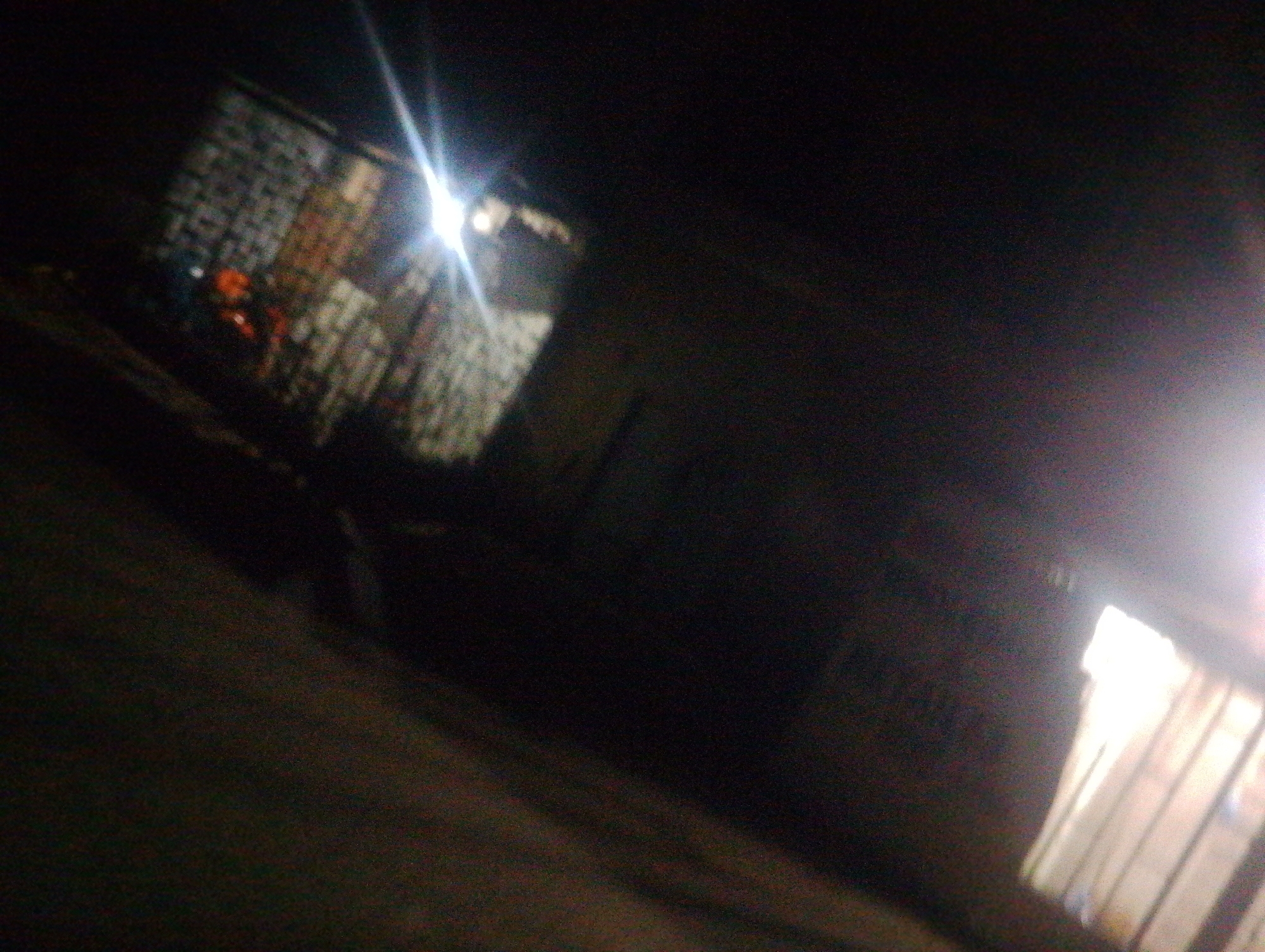 The image depicts a nighttime scene in a dimly lit urban environment, possibly within the city of Jos, Nigeria. The overall quality of the image is poor, characterized by low resolution, blurriness, and significant noise, making detailed identification of specific objects and individuals challenging.
In the left portion of the frame, there appears to be a structure with a textured wall, potentially brick or concrete, exhibiting a grid-like pattern. A bright light source, possibly a streetlamp or a vehicle's headlight, is visible in the upper left, casting a strong glare that obscures details in that area. Faintly visible beneath this glare are hints of color, suggesting the presence of objects or possibly people. One area shows a patch of orange, which could be a piece of clothing or an object.
The right side of the image is dominated by a dark, indistinct background. However, a brightly lit rectangular area, possibly a window or an opening in a building, is prominent. The light emanating from this opening is strong, creating a lens flare effect and rendering the details within it indistinct. The light casts parallel lines onto what appears to be a surface, suggesting the presence of blinds or bars.
The ground in the foreground is dark and untextured, blending into the shadows. The overall impression is of a street or alleyway at night, with limited visibility due to the darkness and artificial lighting. The environmental context suggests a residential or commercial area, typical of urban settings. The poor image quality prevents any definitive conclusions about specific activities or the precise nature of the subjects present.