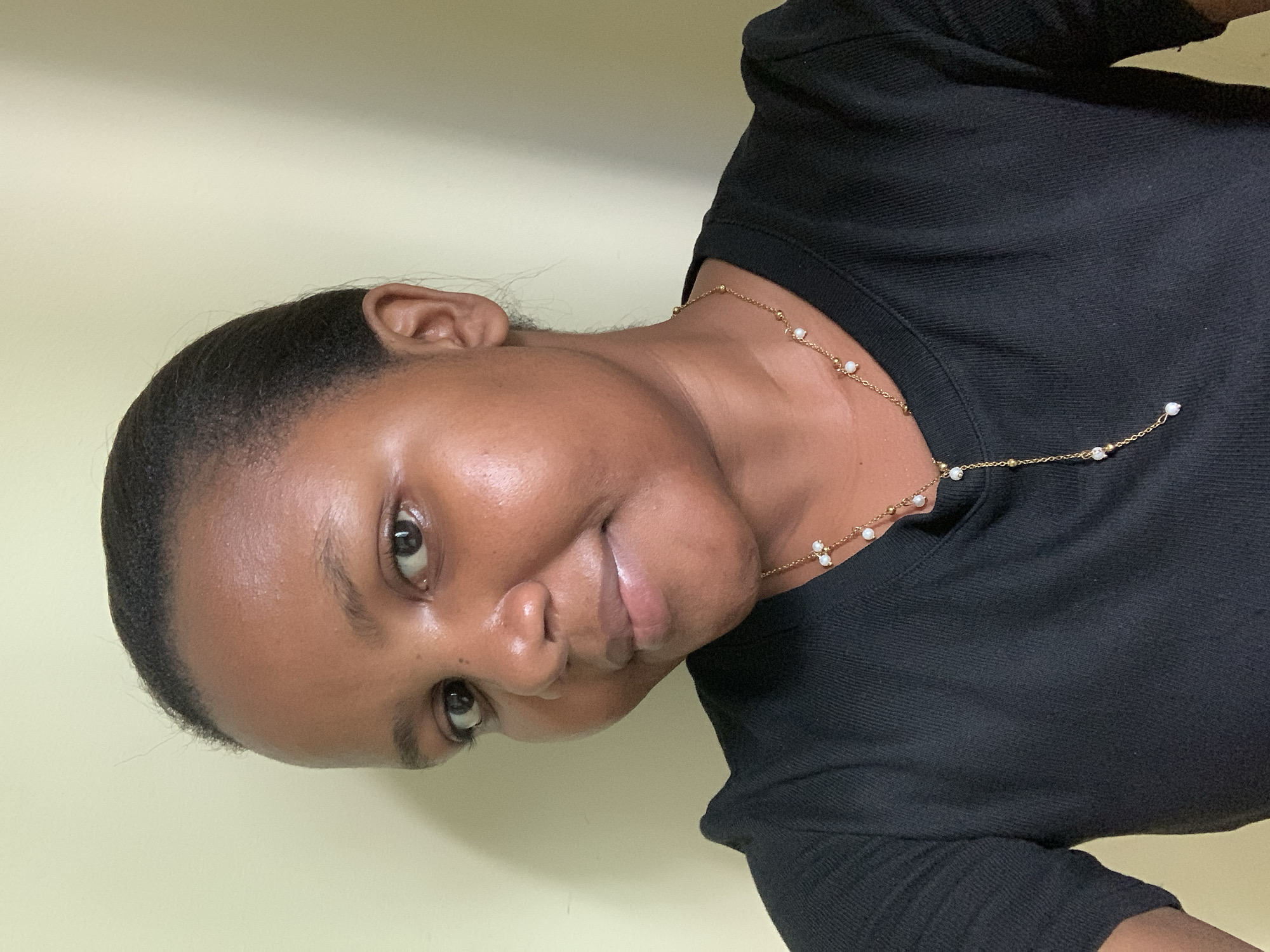 A woman with dark hair and dark skin, wearing a black shirt and a delicate gold necklace with small pearl-like beads, is facing the camera with a slight smile. The background is a plain, light yellow wall. The scene appears to be indoors. The location, Dar es-Salaam, Tanzania, cannot be forensically confirmed from the image alone.
