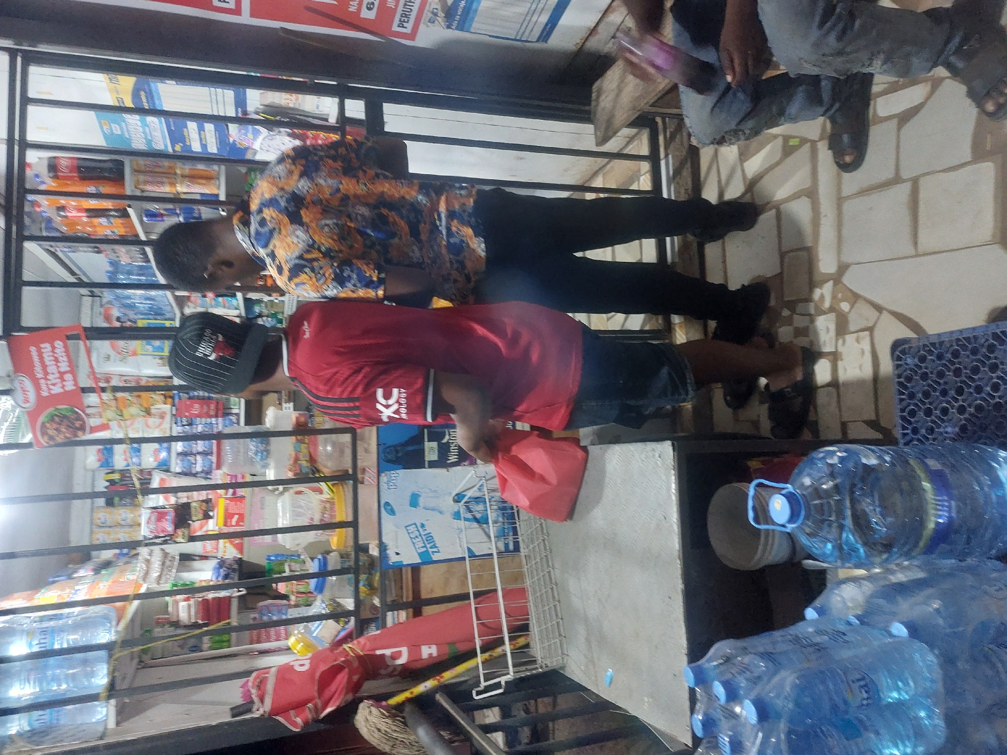 The image shows the front of a small shop in Dar es-Salaam, Tanzania. Two men are present. One is wearing a blue and orange patterned shirt and black pants, looking into the shop. The other man wears a red shirt, a black hat, and shorts. He's holding a red bag, possibly a purchase. The shop is filled with various products, including water bottles, food items, and other goods. Signage with Swahili writing is visible. Outside the shop, there are more water bottles and a mop. The floor is tiled with light-colored stones. Another man is on the far right, visible only from the waist down.