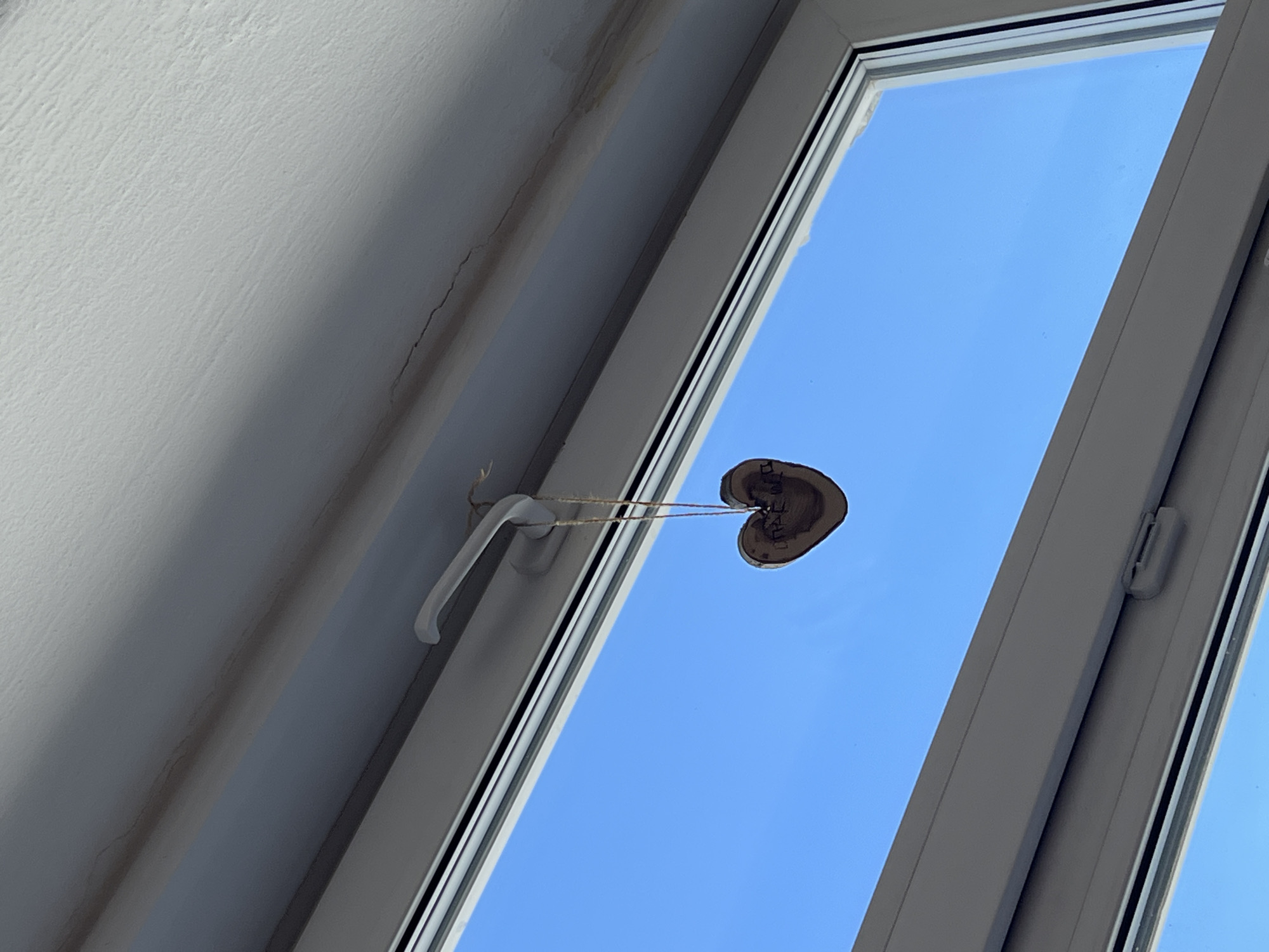 The image depicts an interior scene, likely within a building in Berezniki, Russia, as indicated by the inscription "Березники" on a small, heart-shaped wooden decoration. This decoration hangs from the handle of a white-framed window, suspended by thin, light-colored twine. The window offers a view of a clear, bright blue sky. The interior wall adjacent to the window is painted white and shows some discoloration and minor cracks. The window frame appears to be made of a synthetic material, possibly PVC, and includes standard operating hardware.