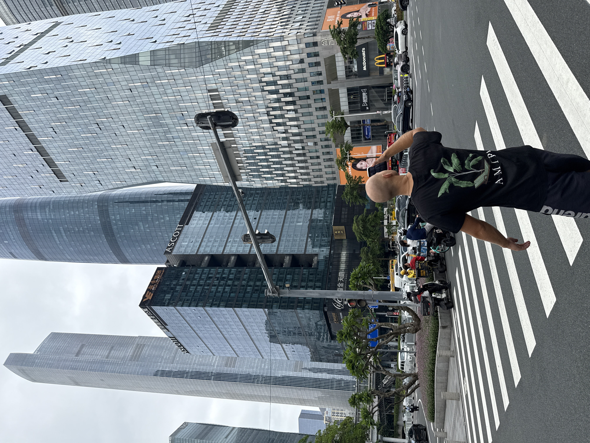 A bald man wearing a black t-shirt with a plant graphic and the word "AMIRI" is standing on a pedestrian crossing. He is holding a smartphone up to his face as if taking a picture or video. Behind him, a street scene unfolds with several modern, glass-fronted skyscrapers, traffic lights, and vehicles. Some of the buildings display corporate logos, including "ASCOTT" and "McDonald's." Trees line the sidewalk, and a traffic light is visible in the foreground. The sky is overcast. The image appears to be taken in Tianhe District, China, known for its contemporary architecture and commercial centers.