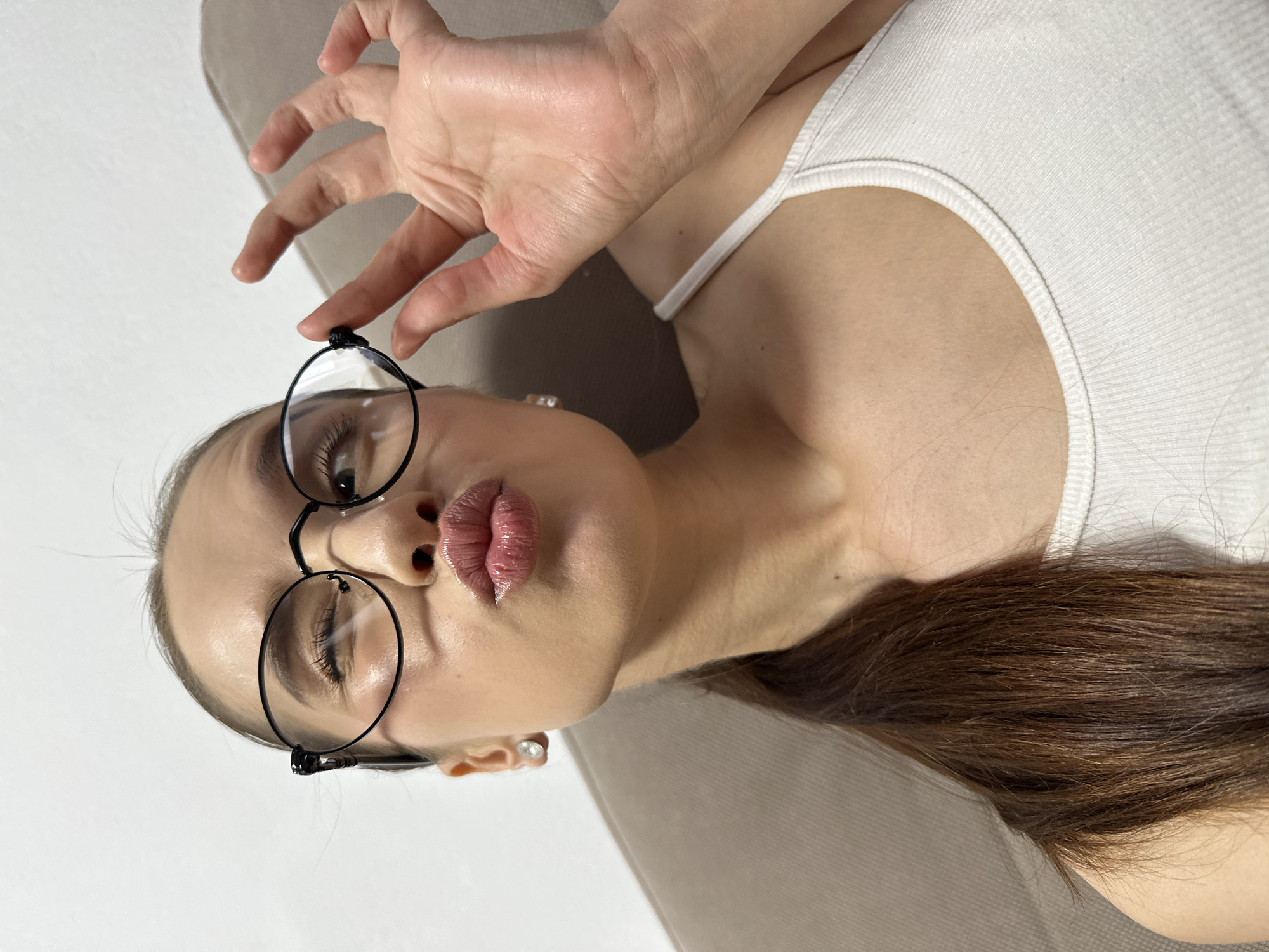 This close-up portrait shows a young woman posing indoors in Yekaterinburg, Russia. She is depicted from the chest up, with her head tilted, appearing to be reclining against a light beige or tan surface, likely a couch or bed, with a plain white wall as the background.
The woman has long, dark brown hair that extends towards the bottom right of the frame. She is wearing a white ribbed tank top. Her prominent lips are pursed in a "kissy face," and she has round, black-rimmed glasses. Her left eye (from her perspective) is closed or significantly squinted, while her right eye is partially visible through the lens. Her right hand is raised, with fingers spread, and her thumb and index finger gently hold the top frame of her glasses near her left temple. She is also wearing small, light-colored stud earrings.
The lighting is soft and diffused, indicating an indoor setting without direct harsh sunlight. The overall atmosphere conveyed by her pose and expression is playful and casual. No text is visible in the image.