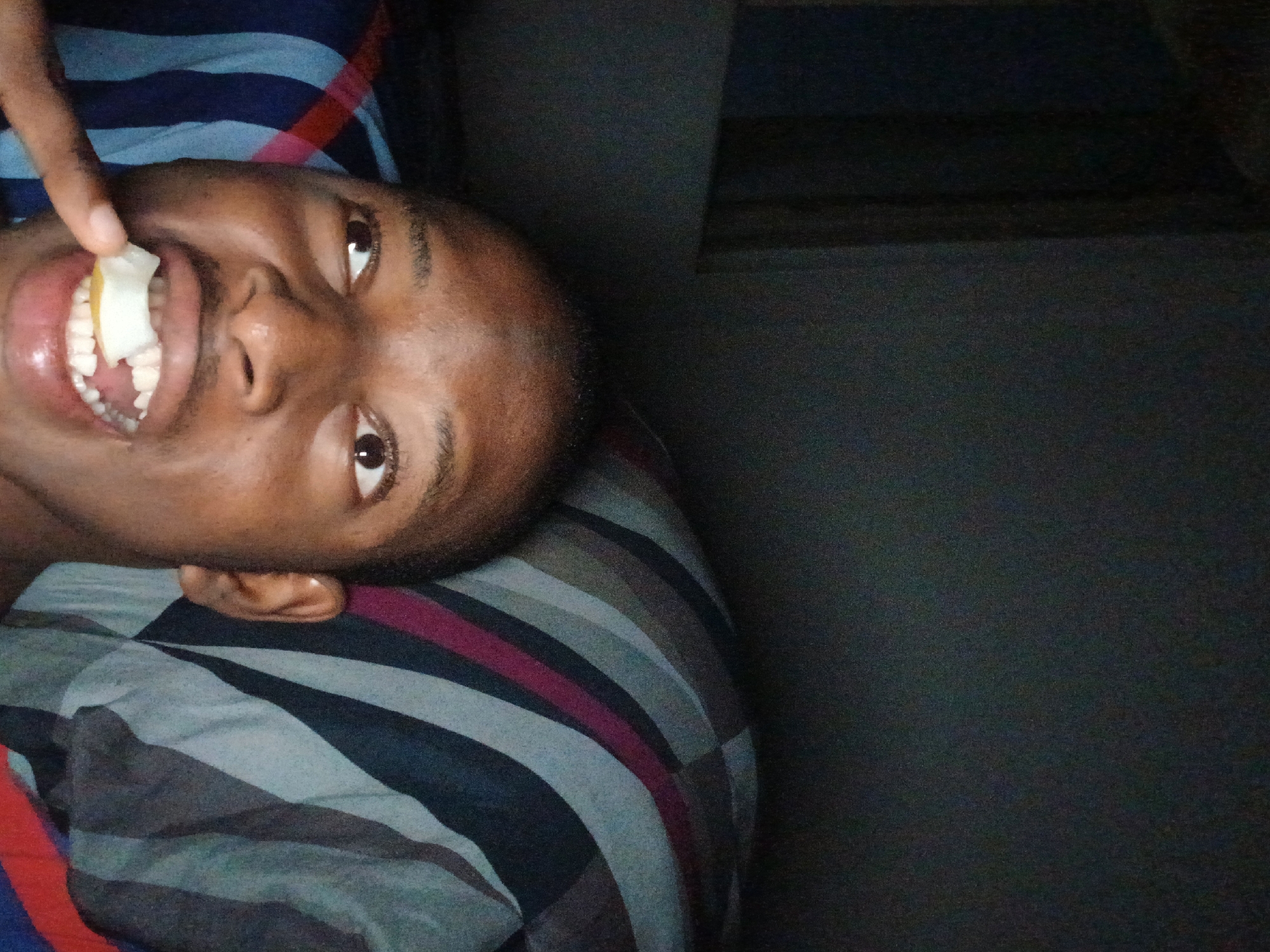 A person is lying on a striped pillow/cushion, holding a white, rectangular object in their mouth with a finger. The person is facing upwards, eyes open, smiling, and looking towards the viewer. The background is dark and out of focus, but suggests a wall or other interior surface. There is no visible information to determine the location of this image in Lagos, Nigeria.