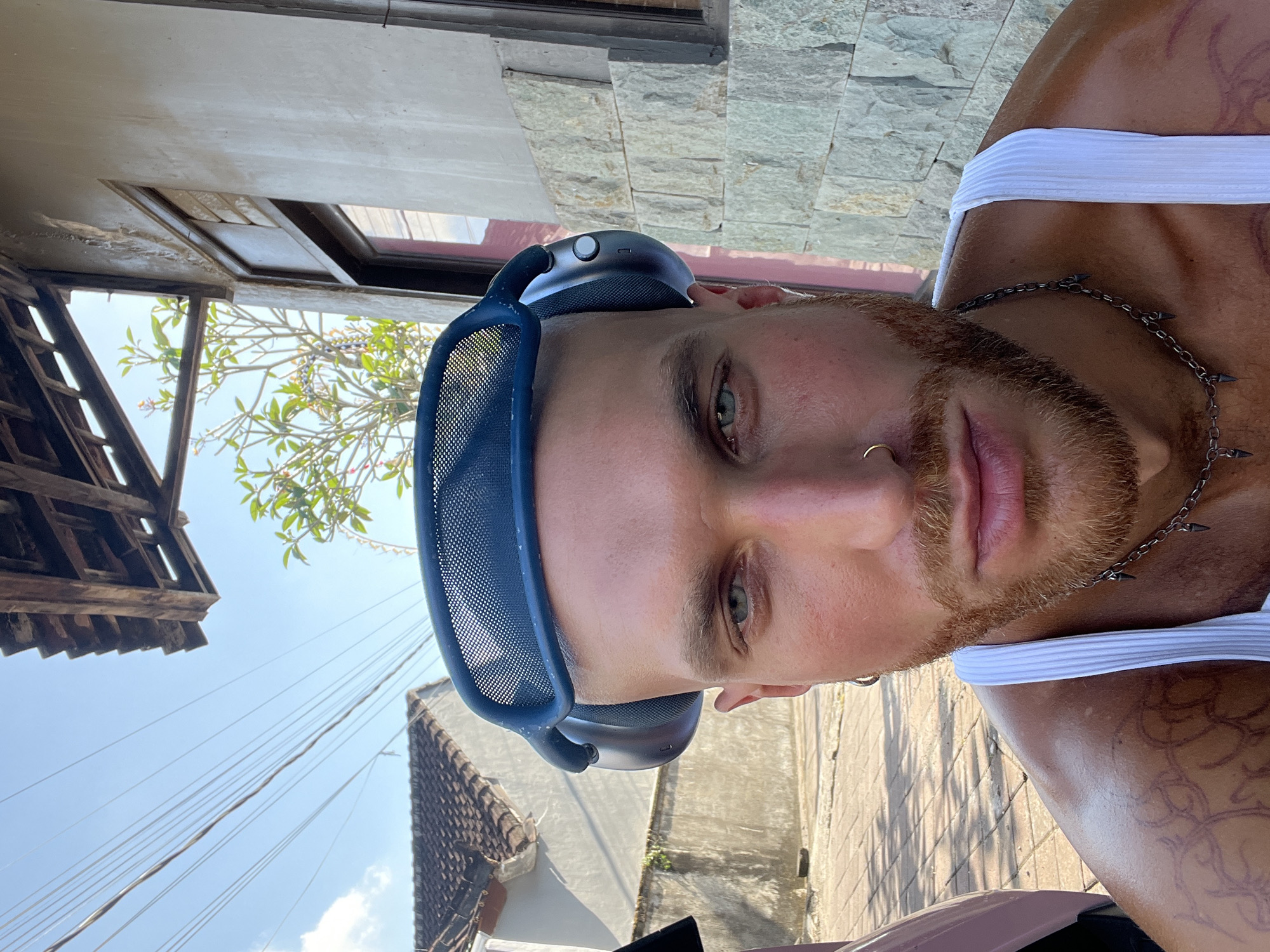 A close-up, waist-up selfie shows a light-skinned man with a shaved head and a red-brown beard wearing large, dark blue headphones with a mesh headband. He is wearing a ribbed white tank top and a silver chain with pointed pendants. A gold hoop earring is in his left ear, and a gold nose ring is in his right nostril. He has tattoos on his left shoulder and upper arm. The man is outdoors, with a cobblestone path in the foreground and a building with a stone facade and a wooden-tiled roof in the background. A tree with green leaves is visible to the right of the man's head. Overhead, power lines stretch across a bright blue sky with a few clouds. The overall setting appears to be a residential area in North Kuta, Indonesia, possibly during the daytime, indicated by the strong sunlight and shadows.