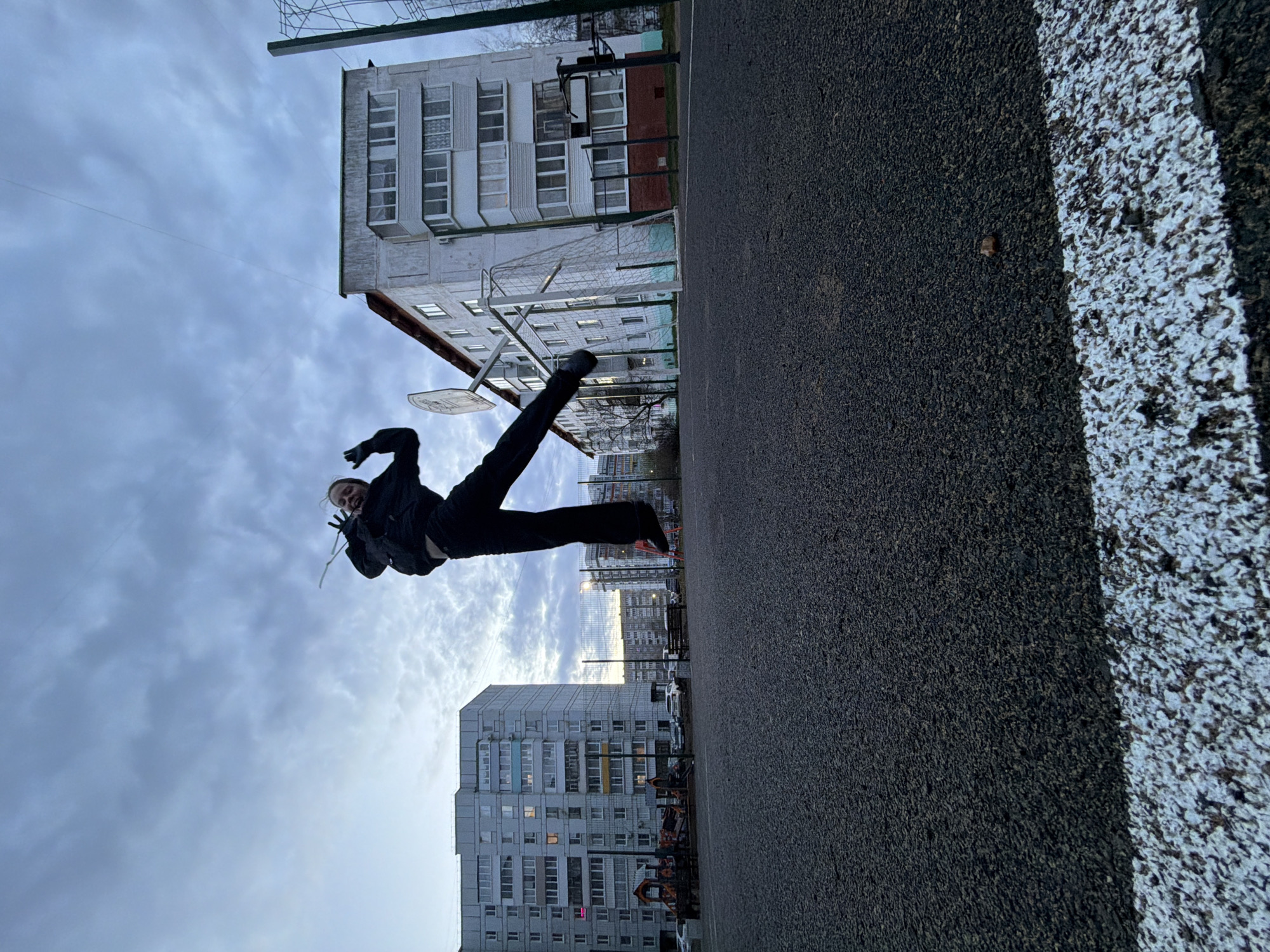 The image captures a dynamic scene on an outdoor asphalt sports court in Chaikovsky, Russia. A person, appearing to be a young individual, is frozen mid-air in a playful jump or acrobatic pose. Dressed in dark clothing – a black jacket, black pants, and black shoes with orange accents – they extend one leg upwards while their arms are bent, and a wide smile is visible on their face, suggesting joy or enthusiasm.
In the background, multiple multi-story residential apartment buildings, typical of Soviet-era architecture, line the horizon under an overcast sky. Some windows in the buildings show lights on, hinting at dusk or early evening. The court itself is made of dark asphalt, with a distinct white painted line visible on the right side, likely marking the boundaries of the playing area.
Behind the jumping person, a metal basketball hoop with a backboard and rim is visible. Further into the background, a white-framed football (soccer) goal with netting is partially seen. A metal mesh fence separates the court from other elements in the distance. The cloudy sky provides soft, even lighting, without strong shadows. No visible text is clearly legible in the image.