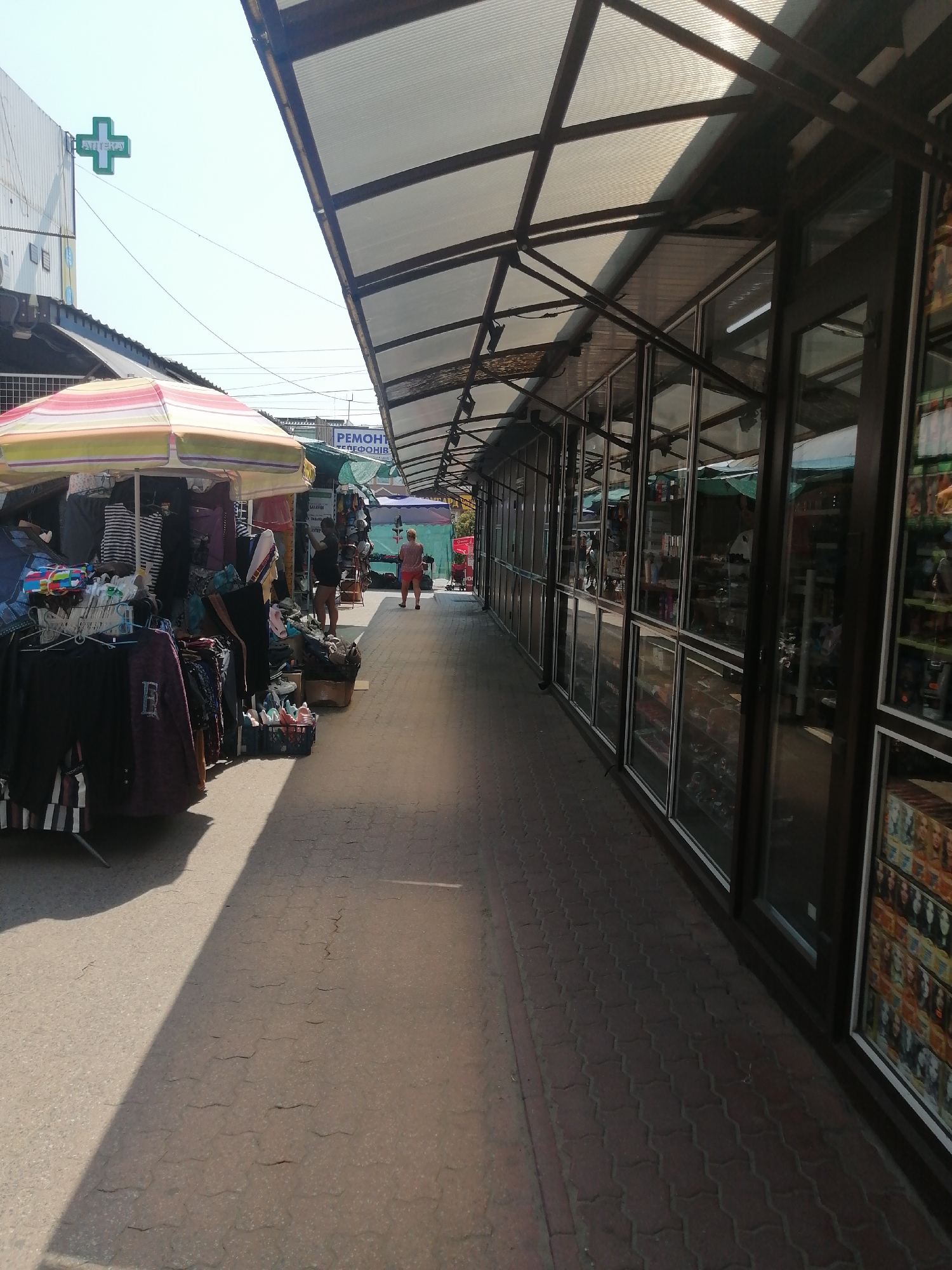 The image depicts an outdoor market scene, likely in the city of Kremenchuk, Ukraine, given the presence of Cyrillic text on signage. In the foreground to the left, a stall displays a variety of clothing items under a striped umbrella. Further down the walkway, a person is seen standing near another stall, and another individual is walking away from the camera. The right side of the image features a row of shops with large glass windows, showcasing merchandise inside. A green pharmacy sign with a cross is visible on a building in the background to the left. The ground is paved with brickwork. The scene is bathed in daylight, with shadows cast by the market structures.