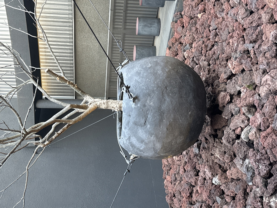 The image displays an outdoor sculptural installation located at Layered Sinsa cafe in Gangnam-gu, Seoul, South Korea. A large, dark grey, spherical planter, approximately 1.5 meters in diameter and exhibiting a textured, weathered surface, is suspended above a ground covered entirely with reddish-brown volcanic rock. A leafless deciduous tree, characterized by a light brown trunk and numerous bare branches, is planted within the suspended vessel. Four black tension cables are visible, attached to metal loops on the planter's rim, extending upwards and outwards to secure the planter approximately 0.5 meters above the volcanic rock bed. In the background, a smooth, dark grey architectural wall is visible on the left. To the right, a lighter grey wall with vertical ribbed panels borders a paved area where three cylindrical dark grey stools, each with a reddish-brown top, are arranged. No individuals or discernible actions are present in the scene.