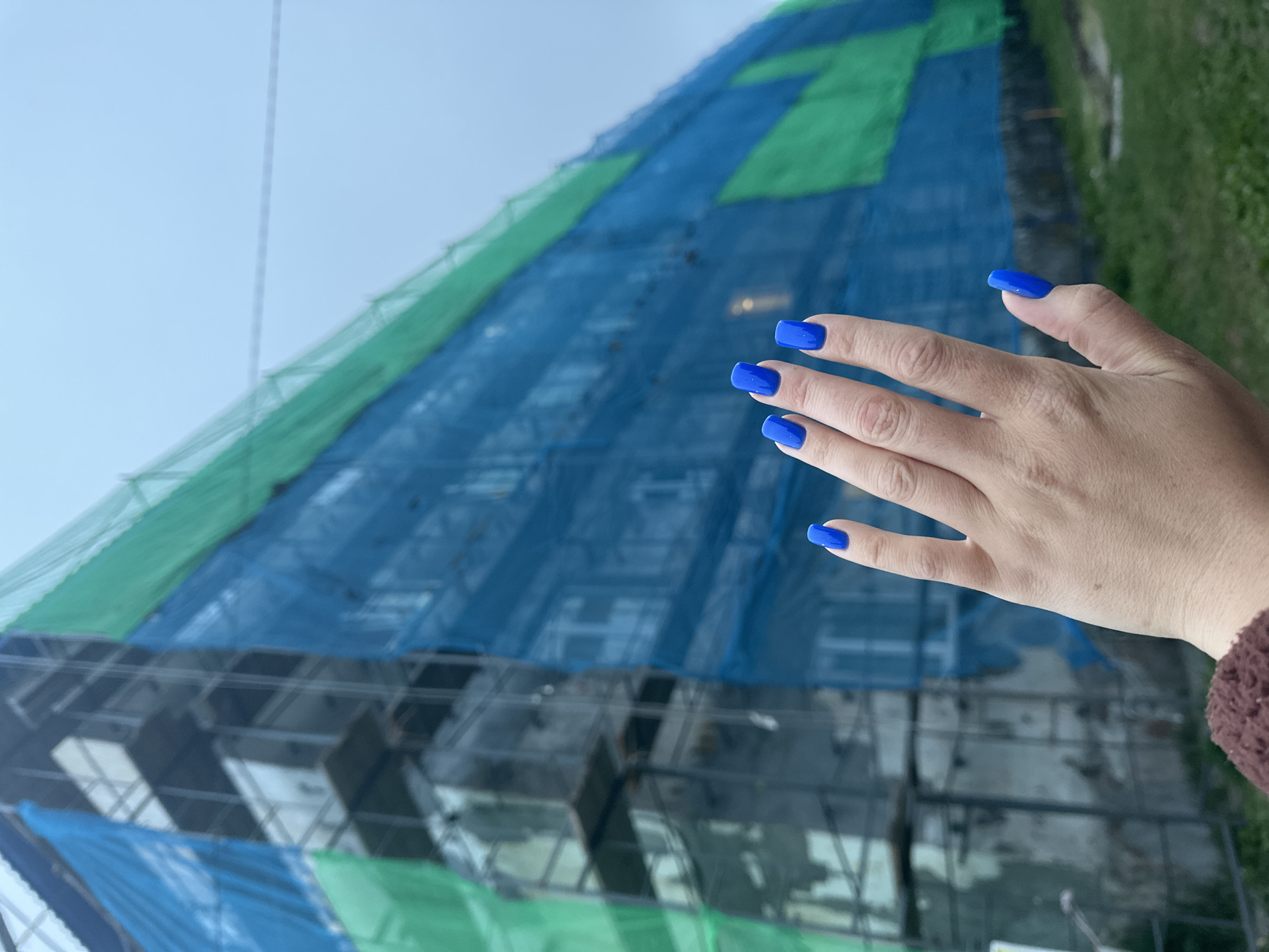 A person's hand with bright blue fingernails is held up in the foreground. Behind the hand, a multi-story building undergoing renovation is covered in scaffolding and green and blue netting. The sky is overcast. The scene is likely a residential building in Vilyuchinsk, Russia, given the context of the request, though the specific location is not visually verifiable within the image itself.