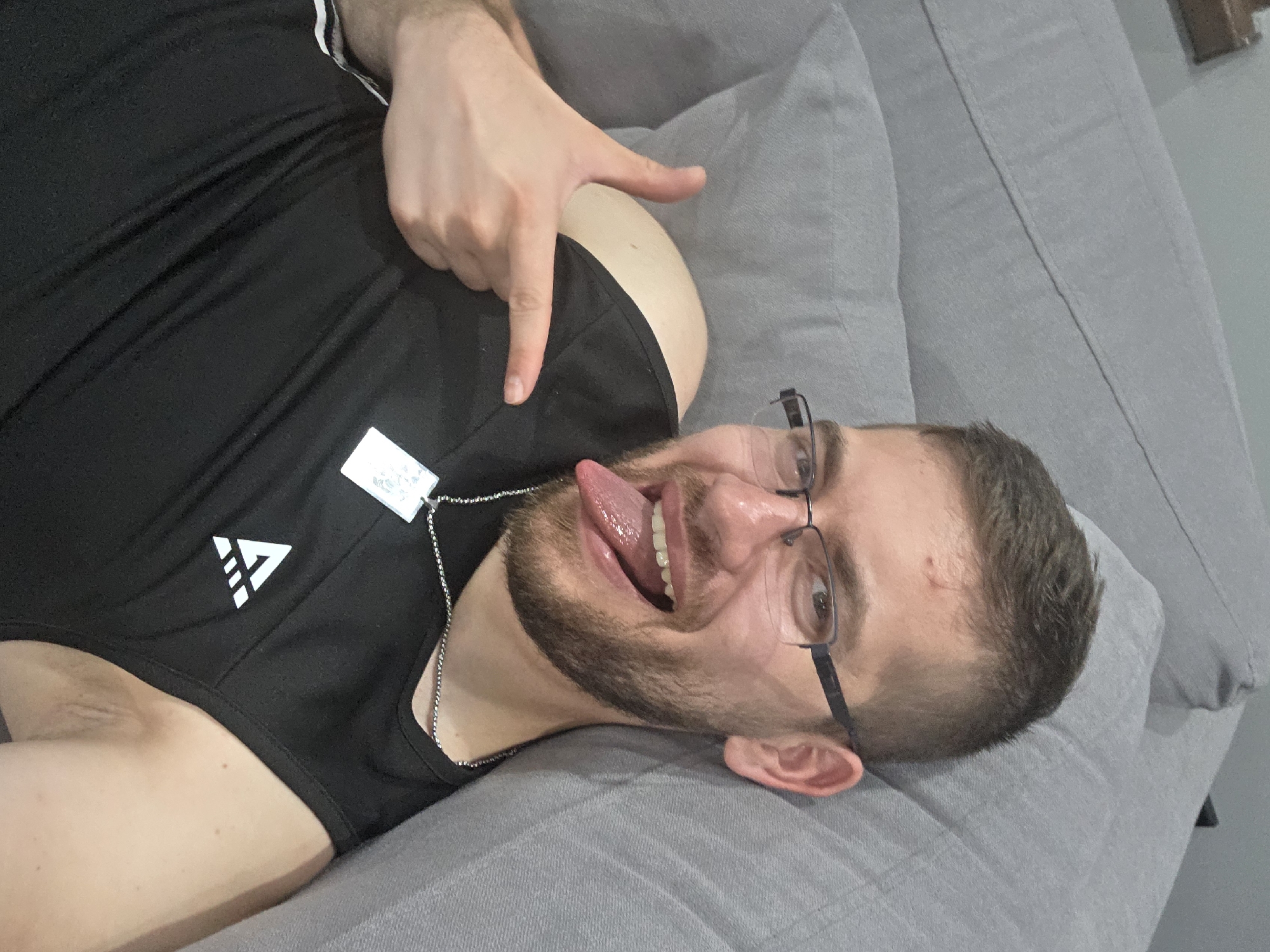 A person is lying on a gray sofa. They are wearing a black tank top and eyeglasses, sticking out their tongue, and making a hand gesture with their right hand. The environment appears to be indoors. There is no information to confirm this picture was taken in Yagodovo, Bulgaria.